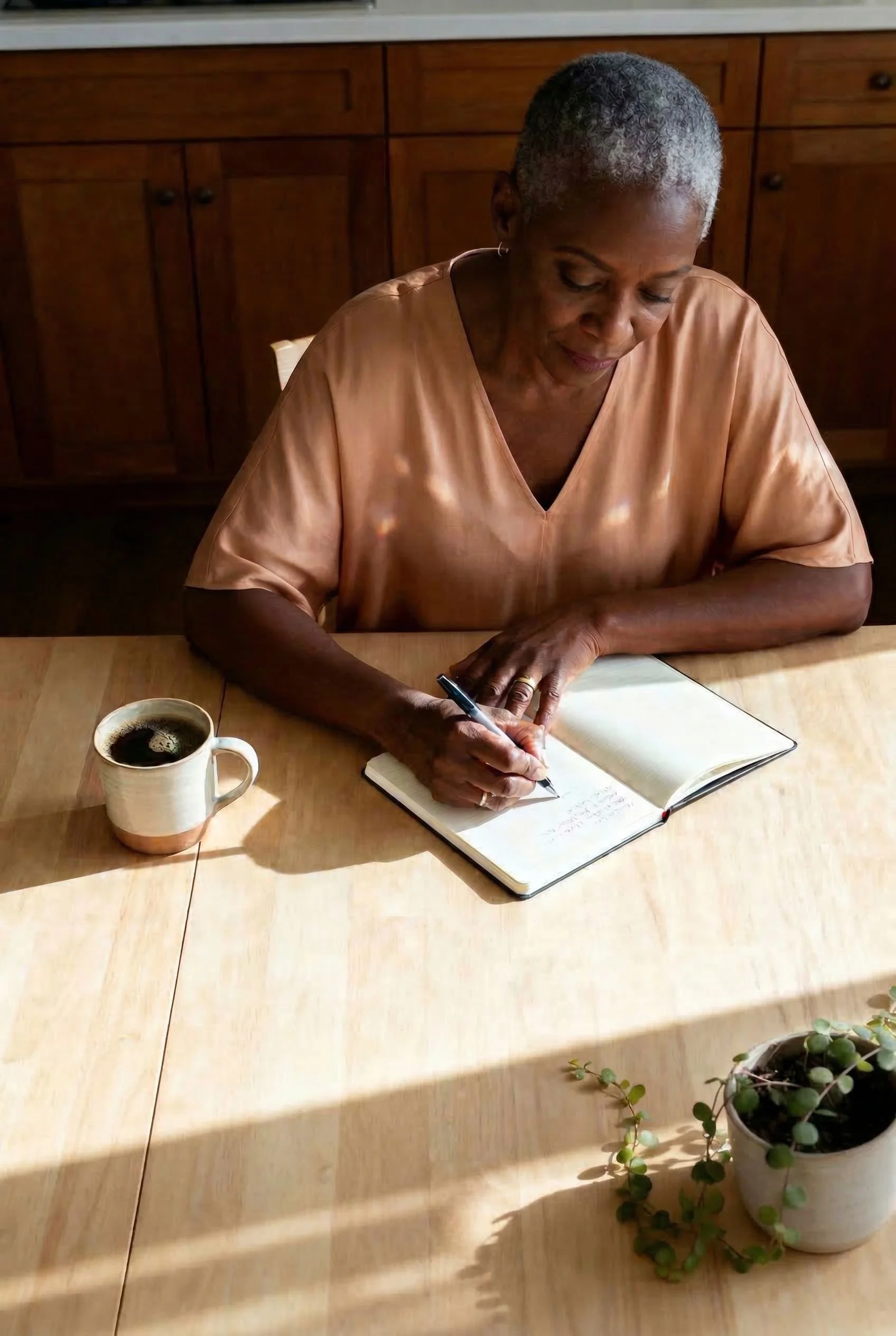 An elderly woman writing in a notebook at a wooden table with a cup of coffee and a potted plant.