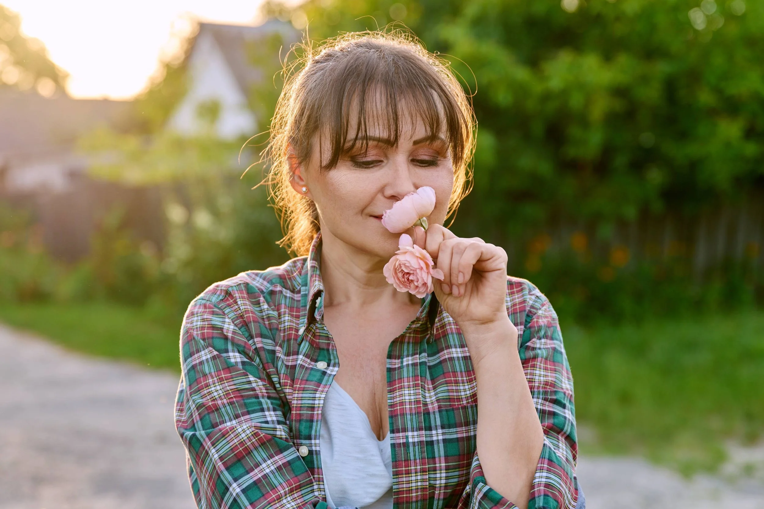 A woman holding pink flowers near her nose with eyes closed outdoors during sunset.
