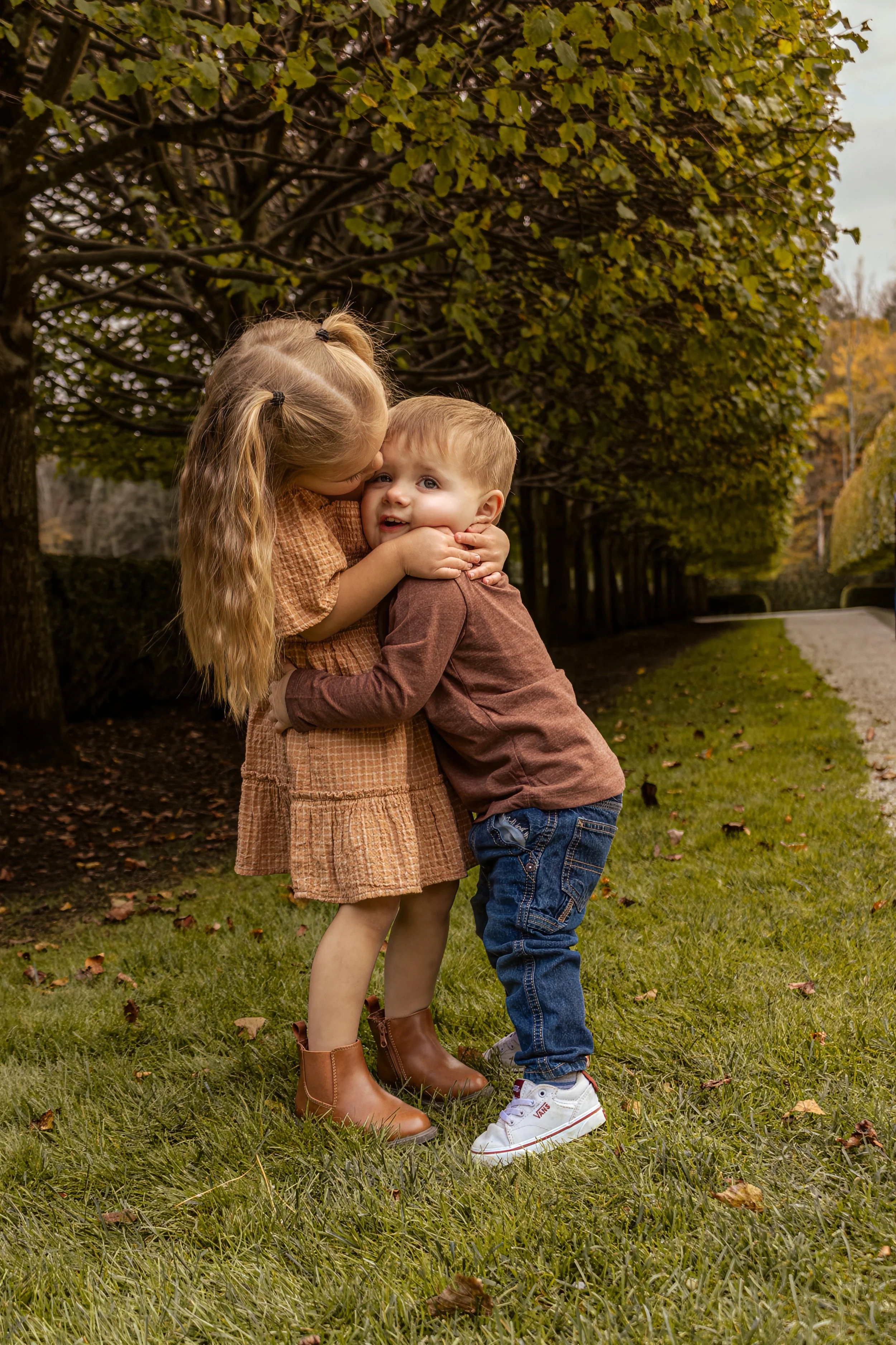 A young girl wearing a brown dress and brown boots hugging a young boy in a brown long-sleeve shirt and blue jeans in a park with a pathway and trees with green leaves.