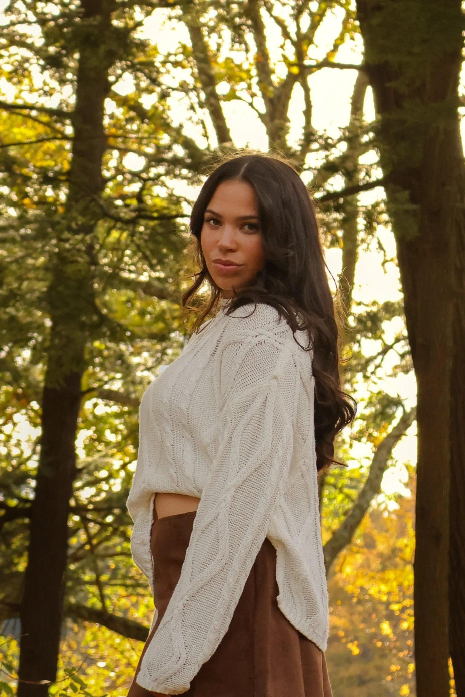 A woman with long wavy dark hair wearing a cream cable knit sweater and brown skirt standing outdoors among trees during fall with yellow and orange leaves.