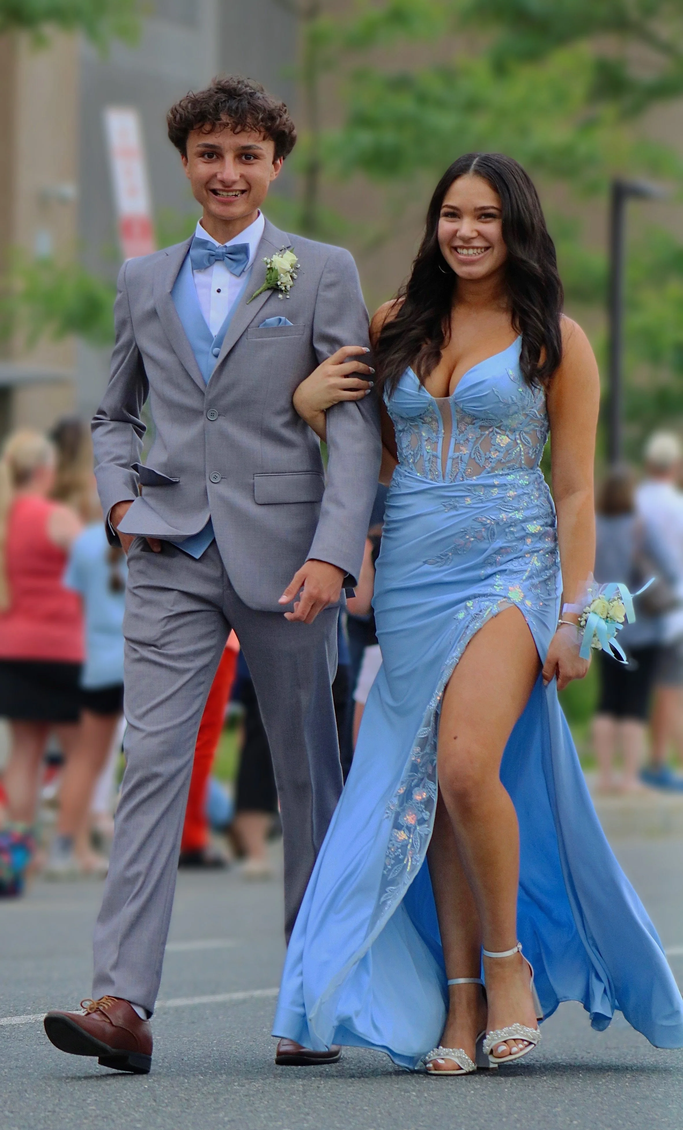 Young couple, dressed in formal attire, walking arm-in-arm at a prom or formal event, with other people in the background.