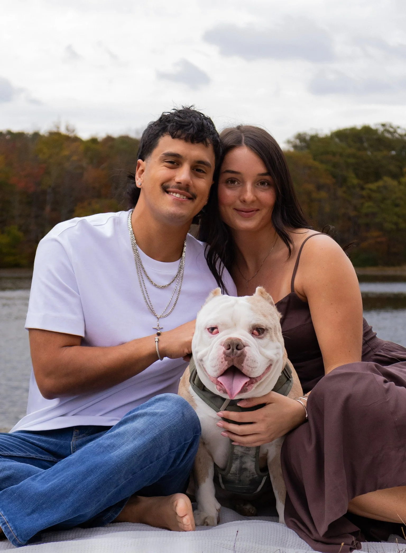 A couple sitting by a lake with their white dog, smiling and enjoying an outdoor moment.