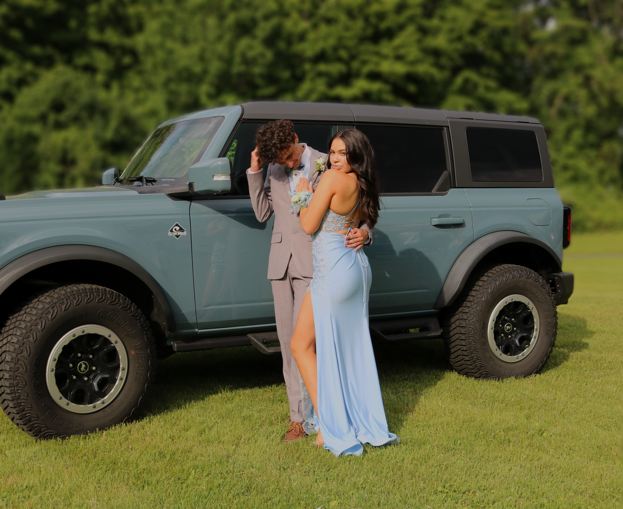 A couple in elegant formal attire standing next to a blue SUV on a grassy field with a blurred green forest background.