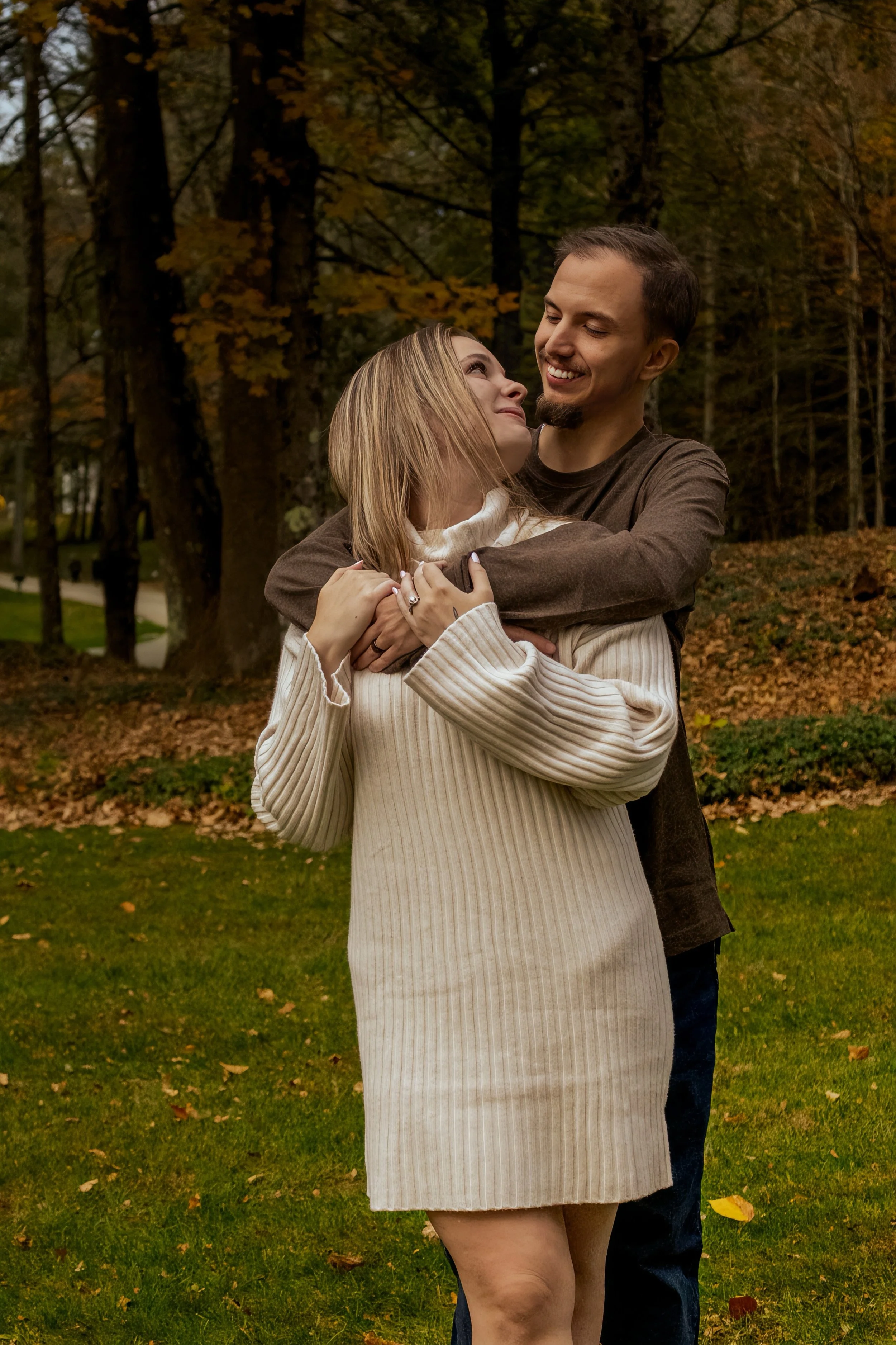 A couple hugging outdoors in a park during autumn, with trees and fallen leaves in the background.