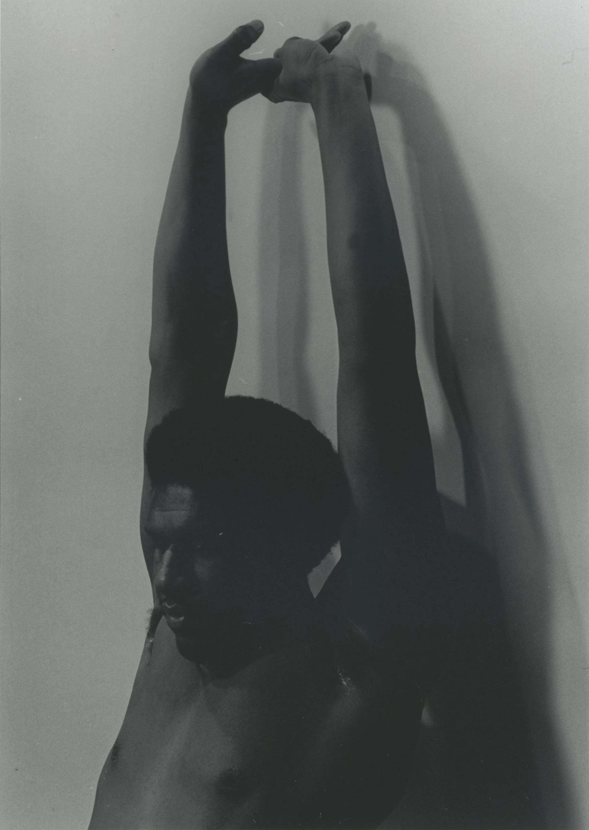 black and white photograph of a Black person, topless and with an afro, looking downward as they stretch their arms to the sky. Their right hand is being held by their left as they stretch.