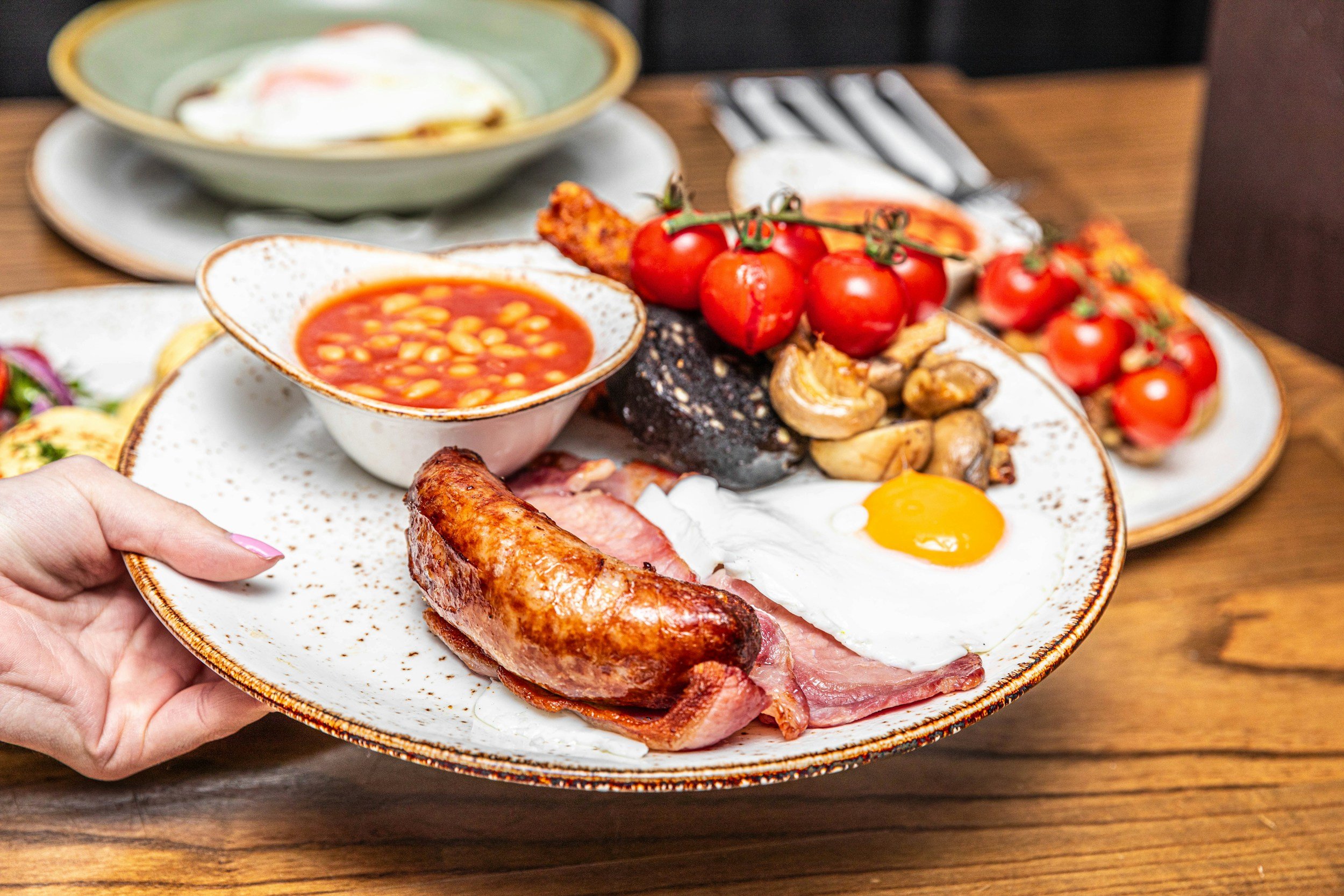A plate of breakfast foods including bacon, a fried egg, a sausage, baked beans, cherry tomatoes, mushrooms, and toast with jam, with a side of baked eggs, in a restaurant setting.
