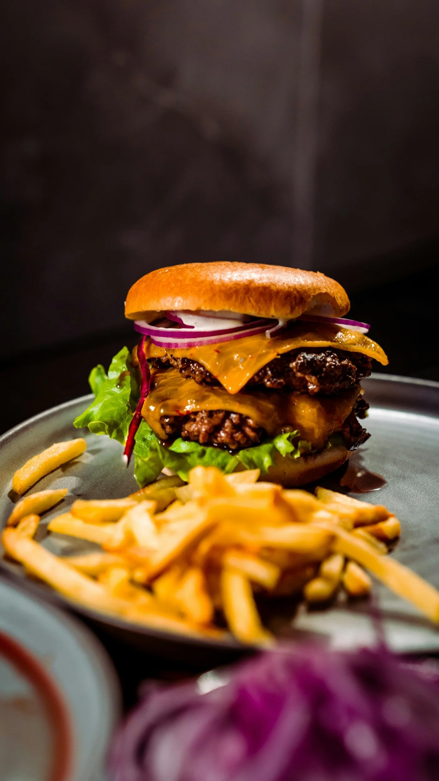 Close-up of a cheeseburger with lettuce, beef patties, melted cheese, pickles, onions, and a bun, served with a side of French fries on a plate.