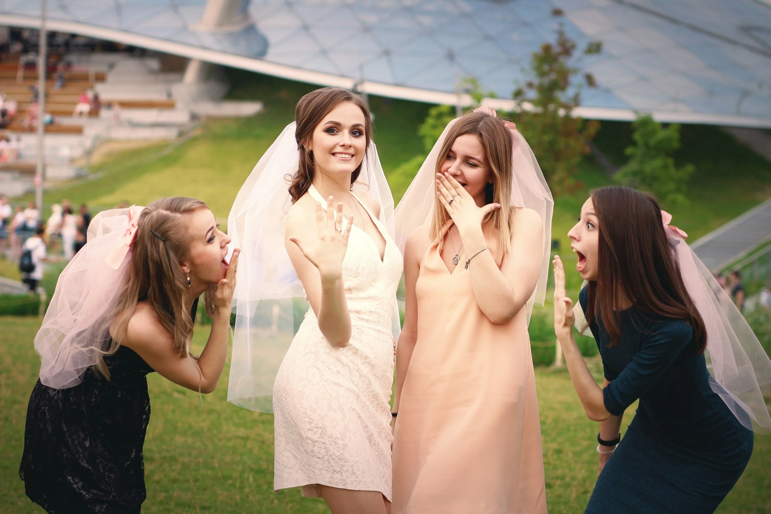 Four women, one dressed as a bride, are celebrating outdoors on a grassy area with a modern building in the background. They are wearing veils and are having an animated, joyful conversation.
