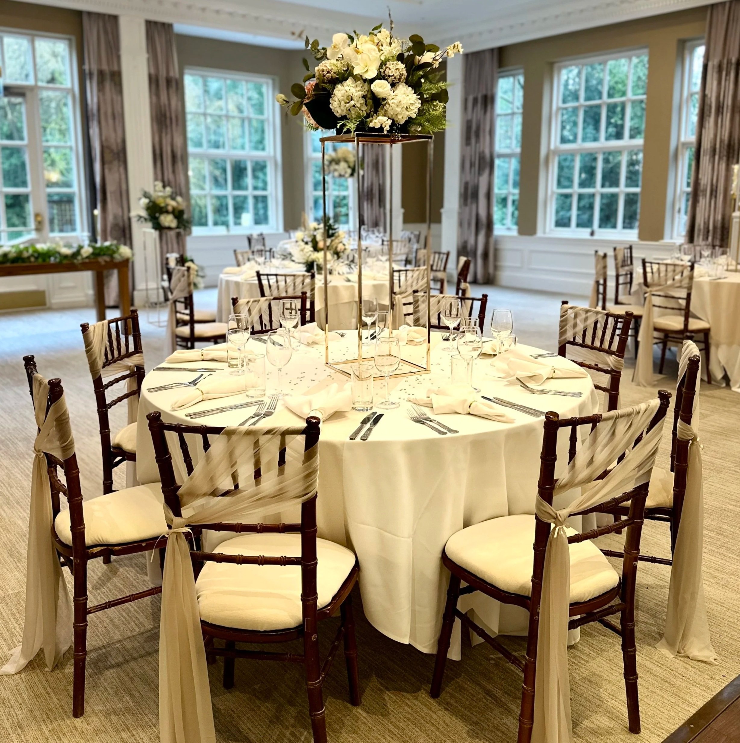 A banquet hall decorated for a wedding or formal event with a round table set with white tablecloth, elegant glassware, and silverware. The table features a tall floral centerpiece with white flowers and greenery. Chivari chairs with white cushions a