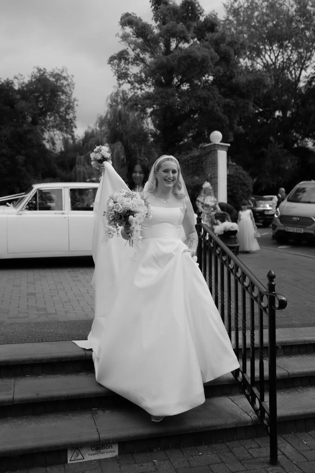 Black and white photo of a bride in a wedding dress, smiling and holding a bouquet, walking down steps outside. Several people and cars are in the background.