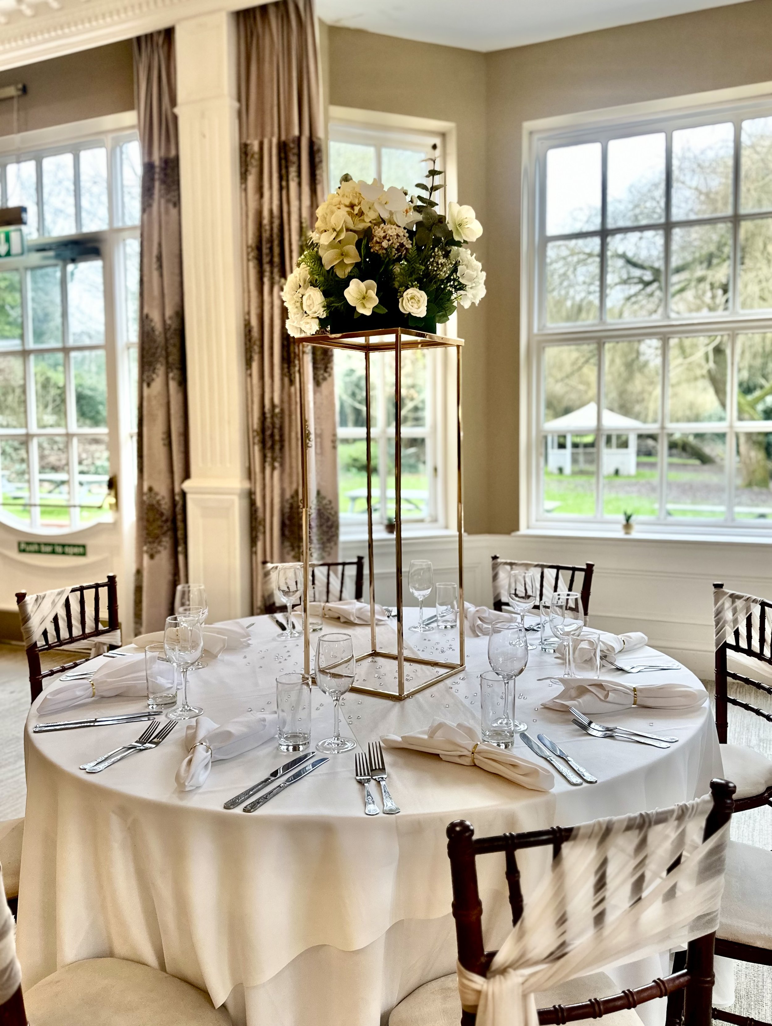 Elegant banquet table with white tablecloth, set for a formal event, featuring a tall floral centerpiece and surrounded by wooden chairs in a bright room with large windows.