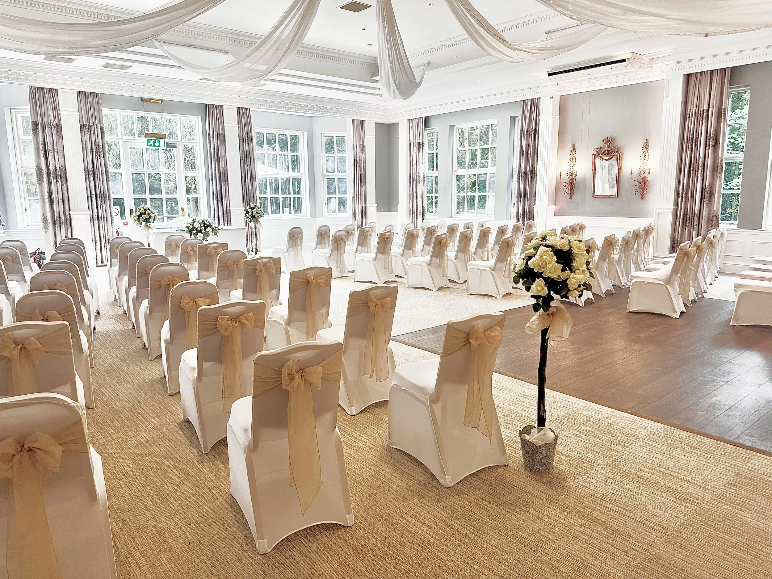 An elegant wedding ceremony setup in a bright, spacious room with white chairs decorated with beige covers and bows, arranged in rows facing a small platform with a floral arrangement. The room has large windows, draped curtains, and decorative ceili