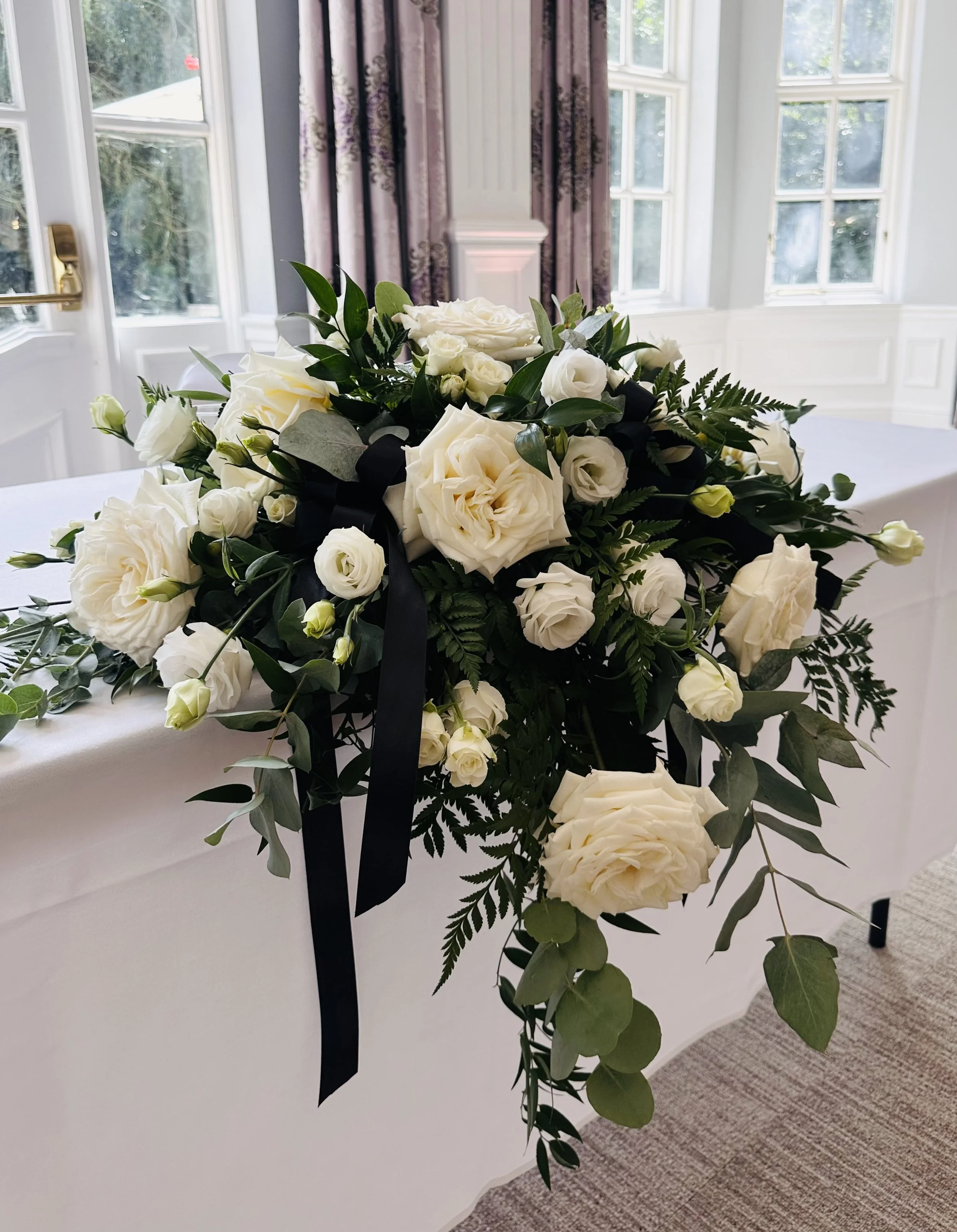 White floral arrangement with roses and greenery on a white table in a bright room with windows and curtains.