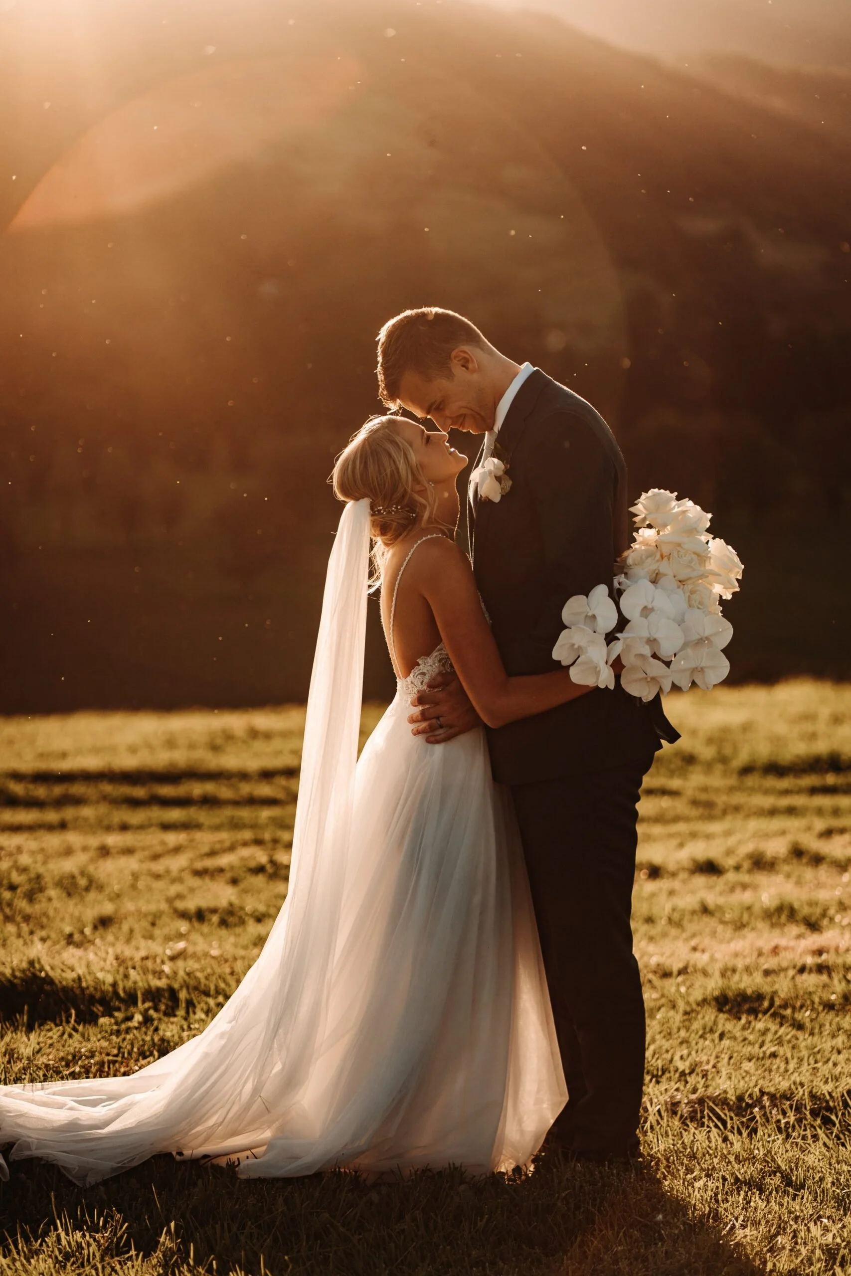A bride and groom share an intimate moment outdoors during sunset, with the bride holding a bouquet of white flowers, both smiling and leaning their foreheads together.