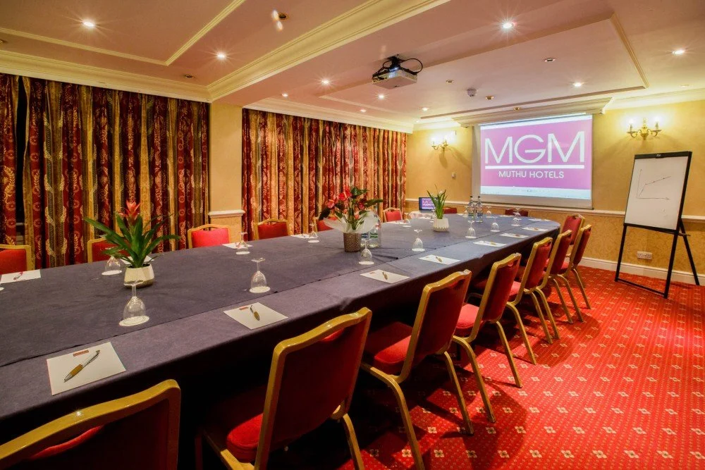 Meeting room set up with a long table, chairs, potted plants, and a presentation screen displaying the MGM Muthu Hotels logo.
