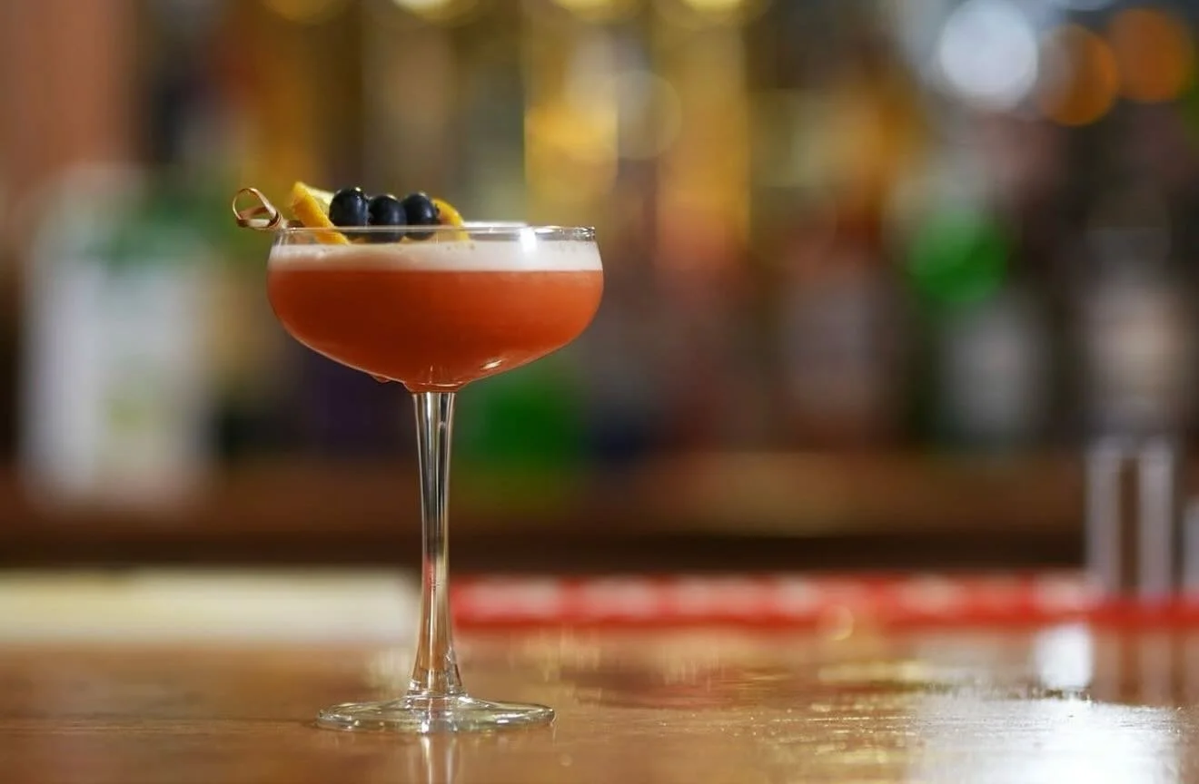 A cocktail glass with a pink-colored drink garnished with blueberries and lemon peel