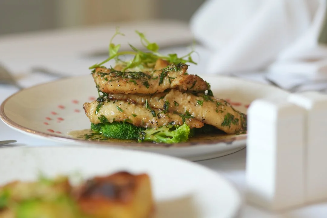 A plate of grilled fish topped with herbs, garnished with microgreens, sitting on a table with white tablecloth and napkins.