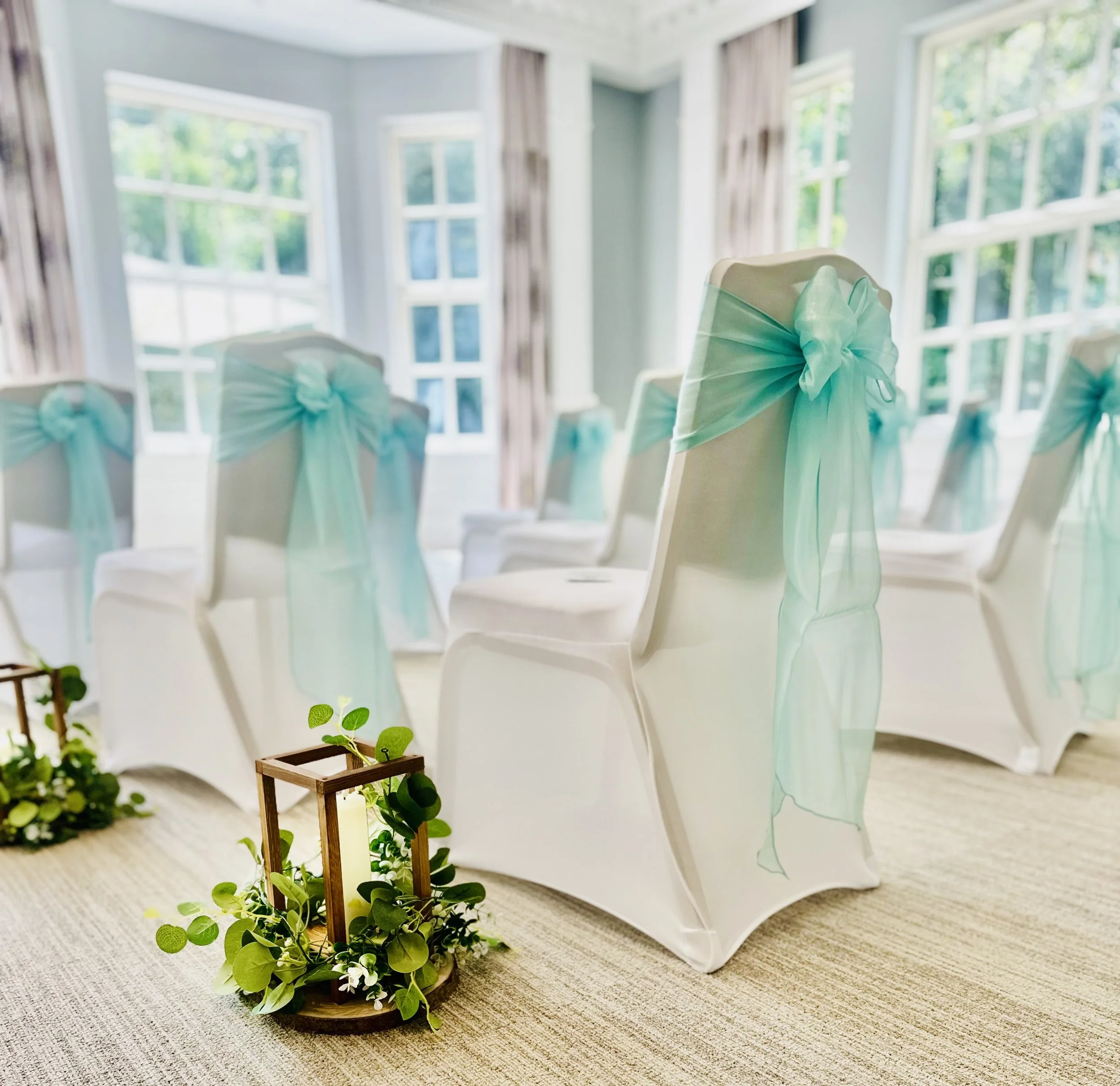 Chairs decorated with light blue tulle bows for a wedding ceremony inside a bright room with large windows.