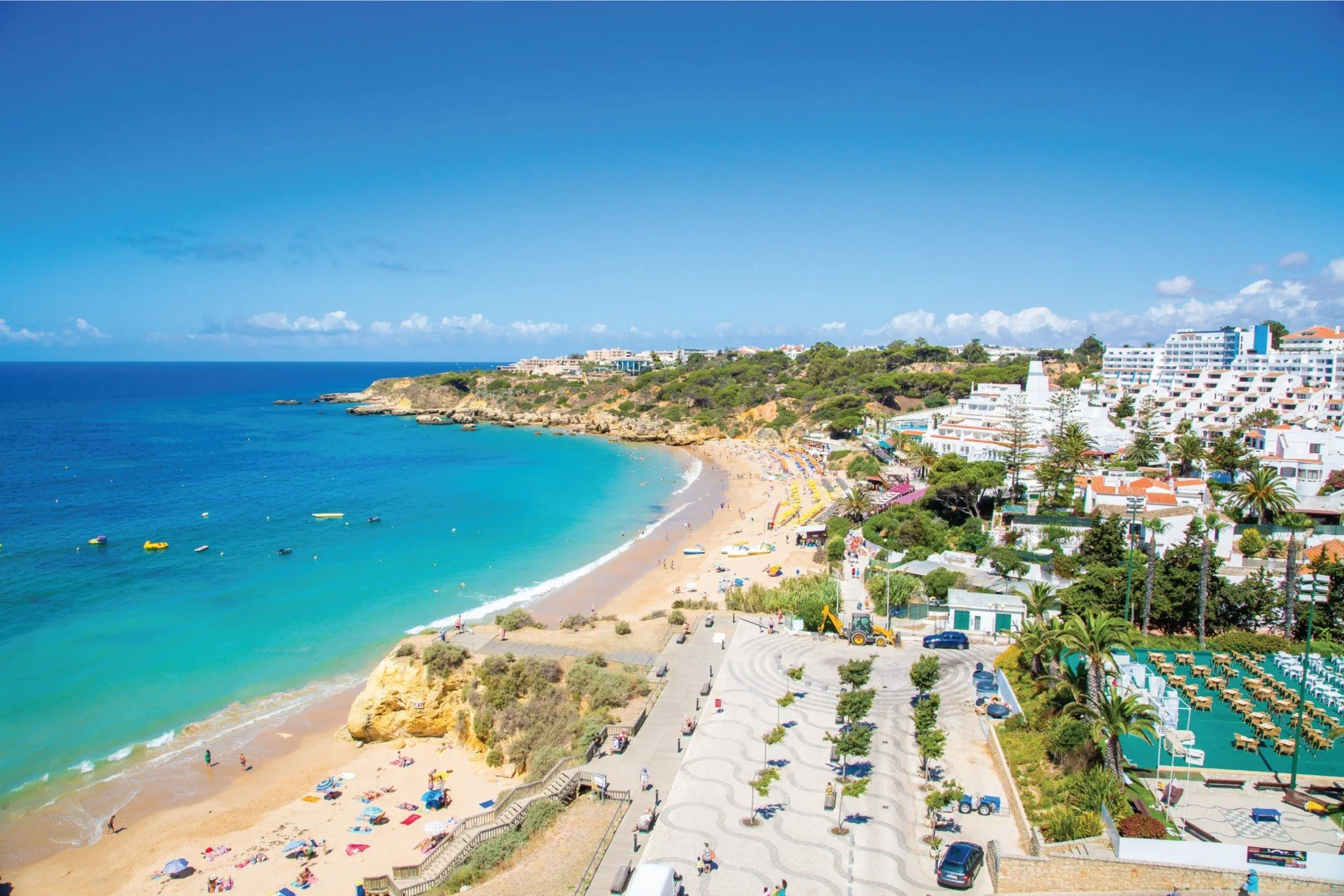 Aerial view of a tropical beach with turquoise water, sandy shore with umbrellas, and a promenade with palm trees and parked cars, with white buildings and lush green vegetation in the background under a clear blue sky.