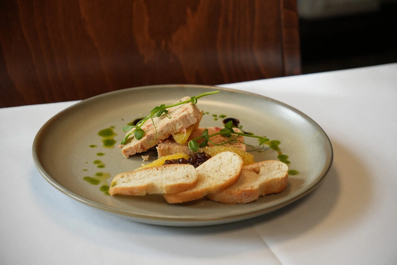 Slices of bread with a terrine, garnished with microgreens, accompanied by yellow and dark fruit preserves, and drizzled with green herb oil on a neutral-colored plate.