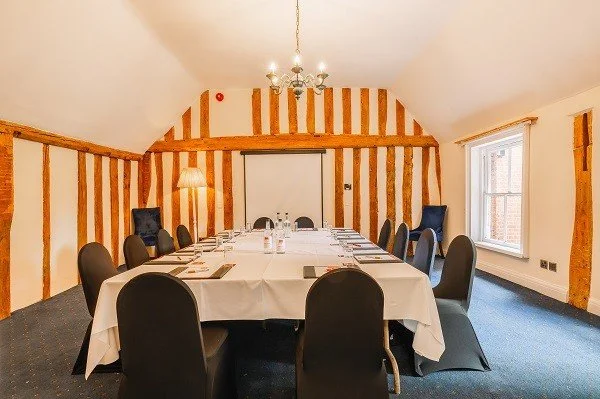 Conference room with a long table covered with a white tablecloth, surrounded by black chairs, with a window on the right and wooden wall accents, chandelier hanging from the ceiling.