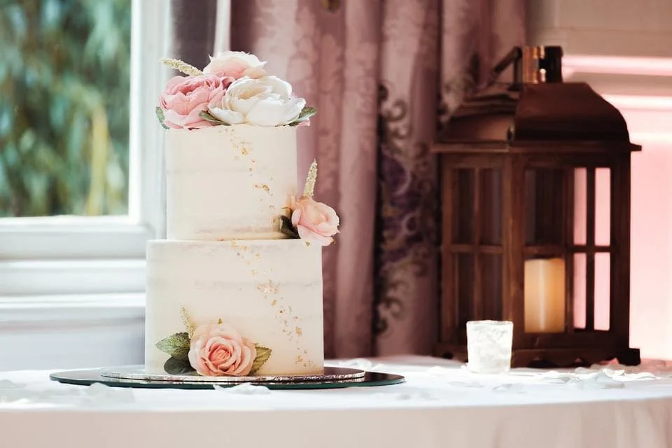 Two-tiered wedding cake decorated with pink and white flowers, gold accents, and greenery, placed on a silver tray on a table with a white tablecloth, with a lantern and window in the background.