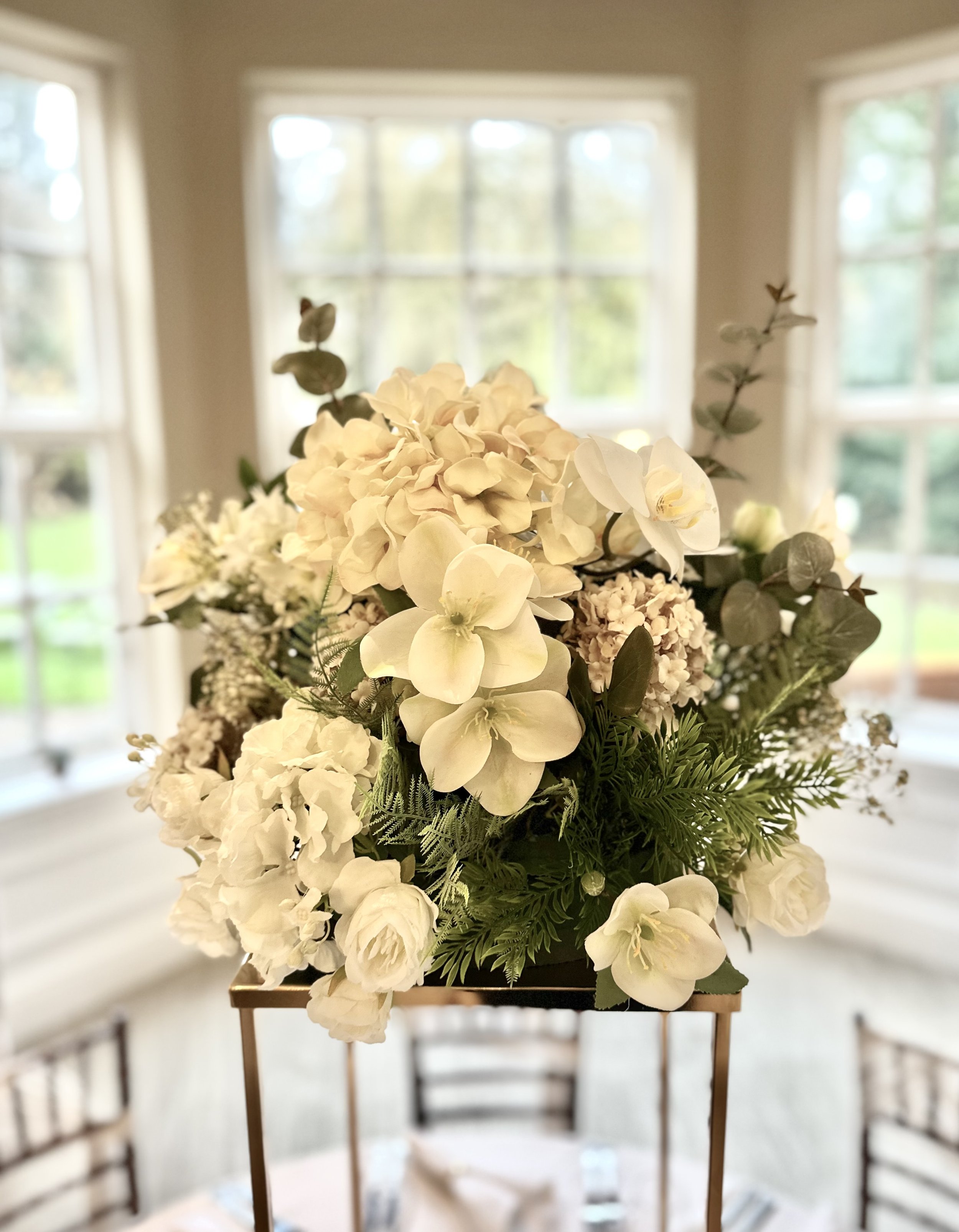 A floral arrangement of white flowers including hydrangeas and roses on a table in a room with large windows and a green outdoor view.