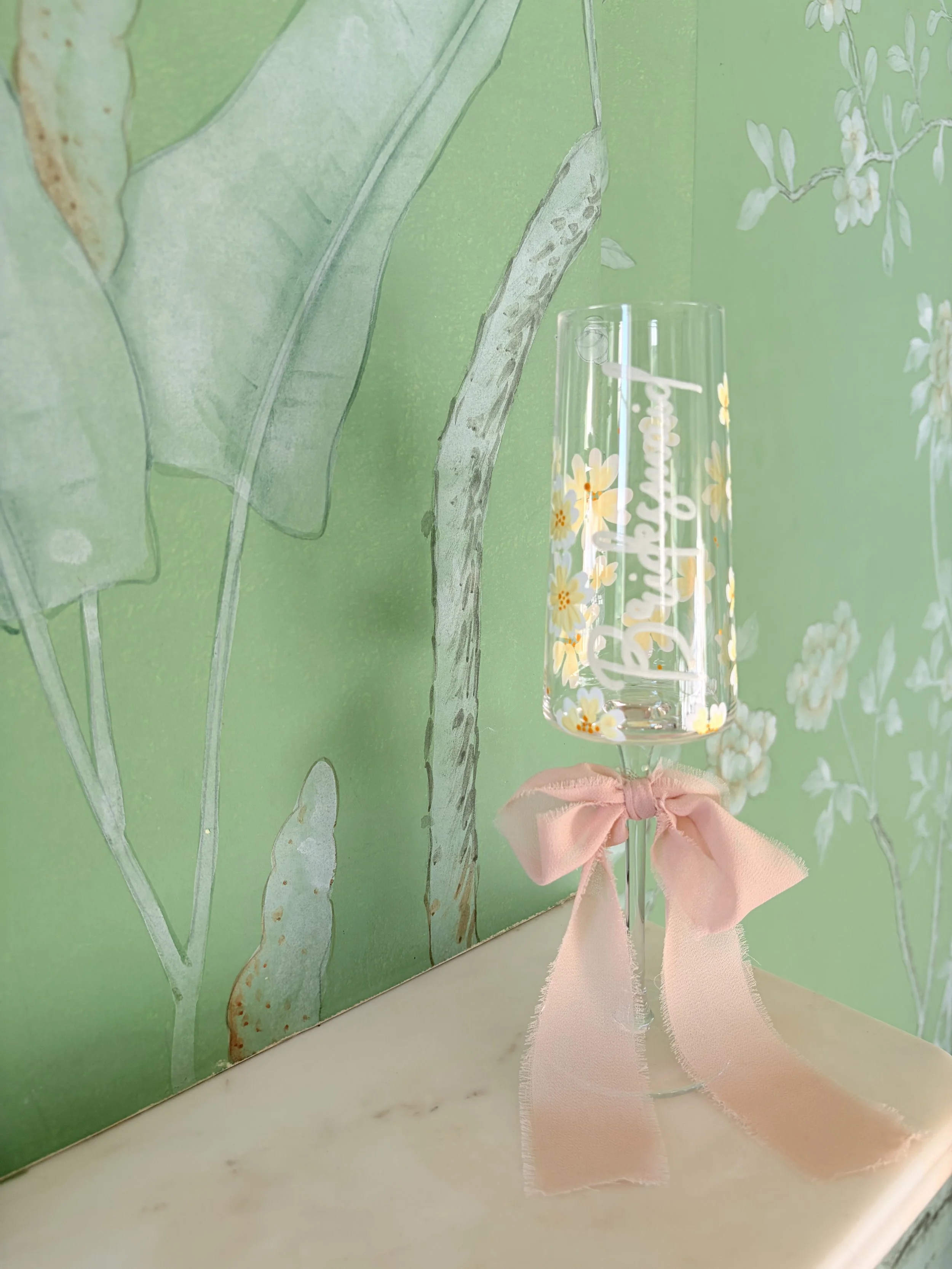A clear champagne flute with floral designs and the word 'Celebration' written on it, decorated with a pink ribbon bow, placed on a white marble surface against a green leafy wallpaper background.
