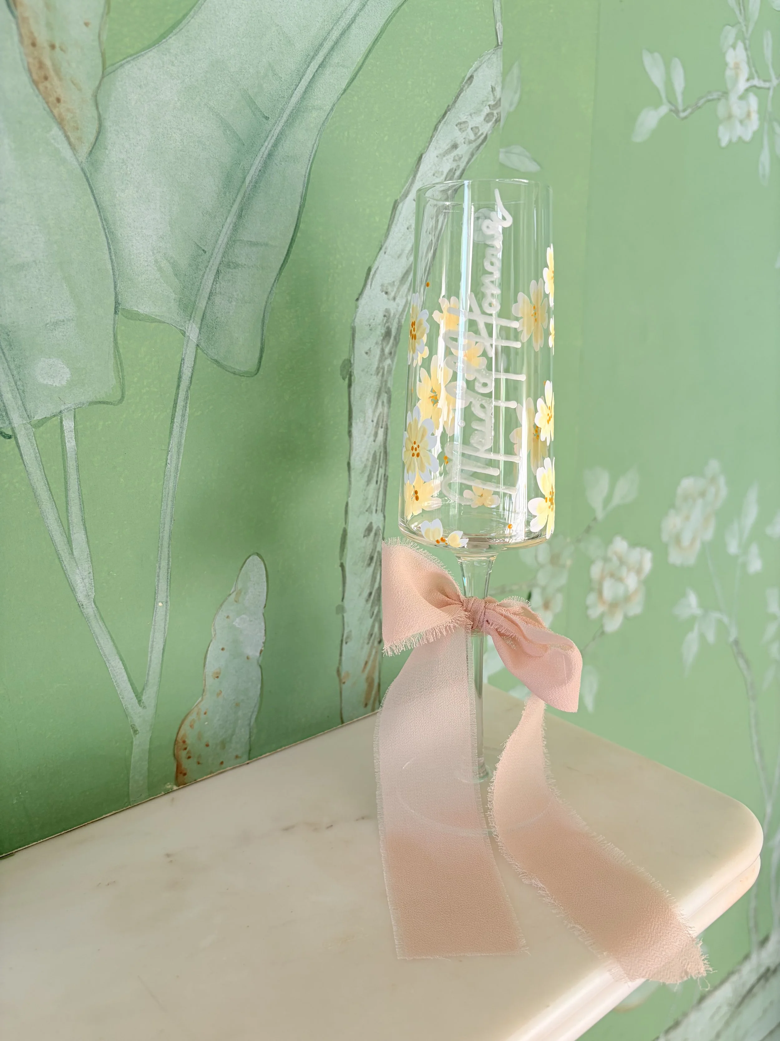 Champagne flute with floral design and pink ribbon, placed on a marble surface against a green floral wallpaper background.