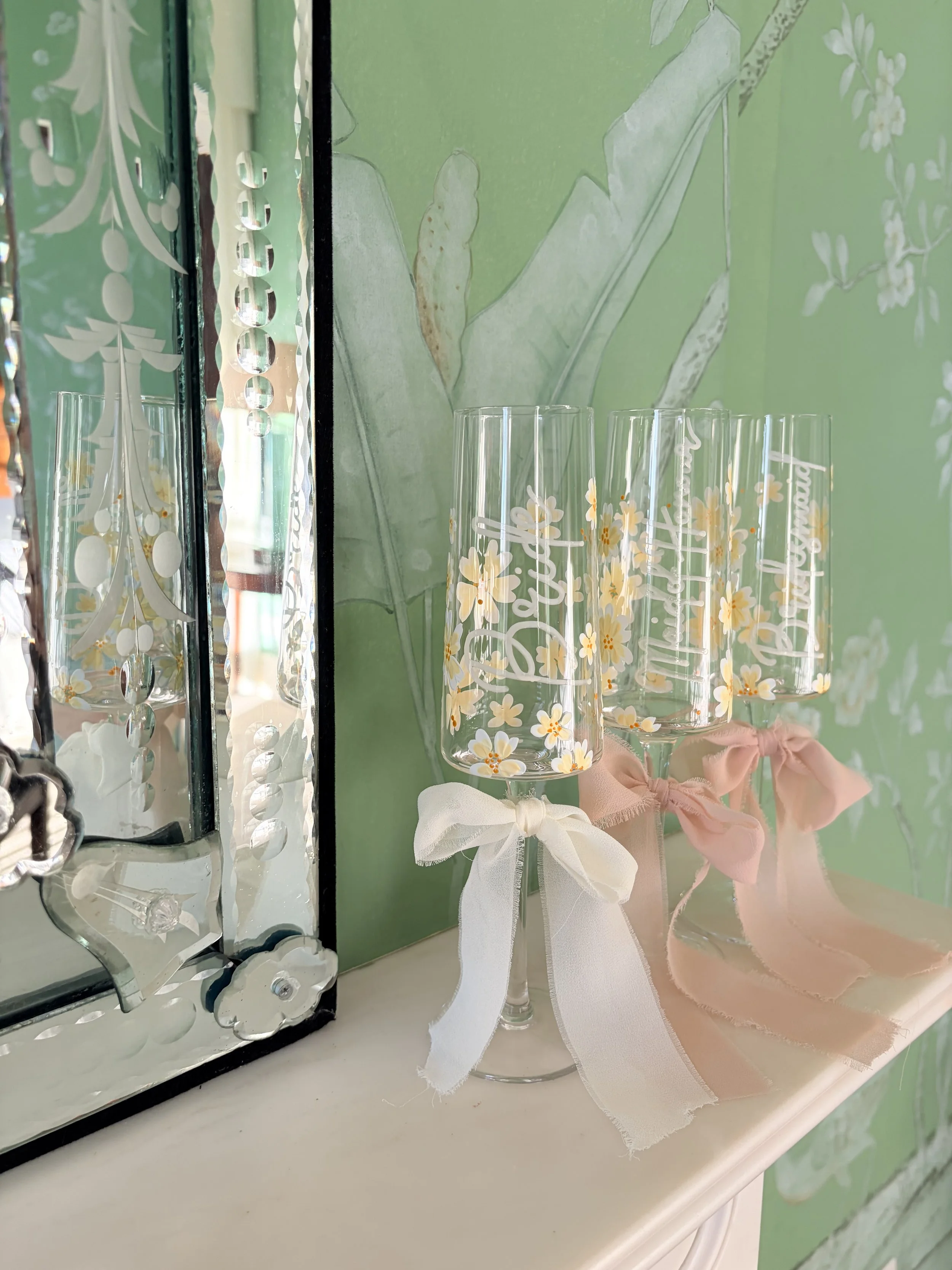 Three tall champagne flutes decorated with white and yellow daisy patterns, each tied with a white or pink ribbon, placed on a white surface against a green floral wallpaper background.