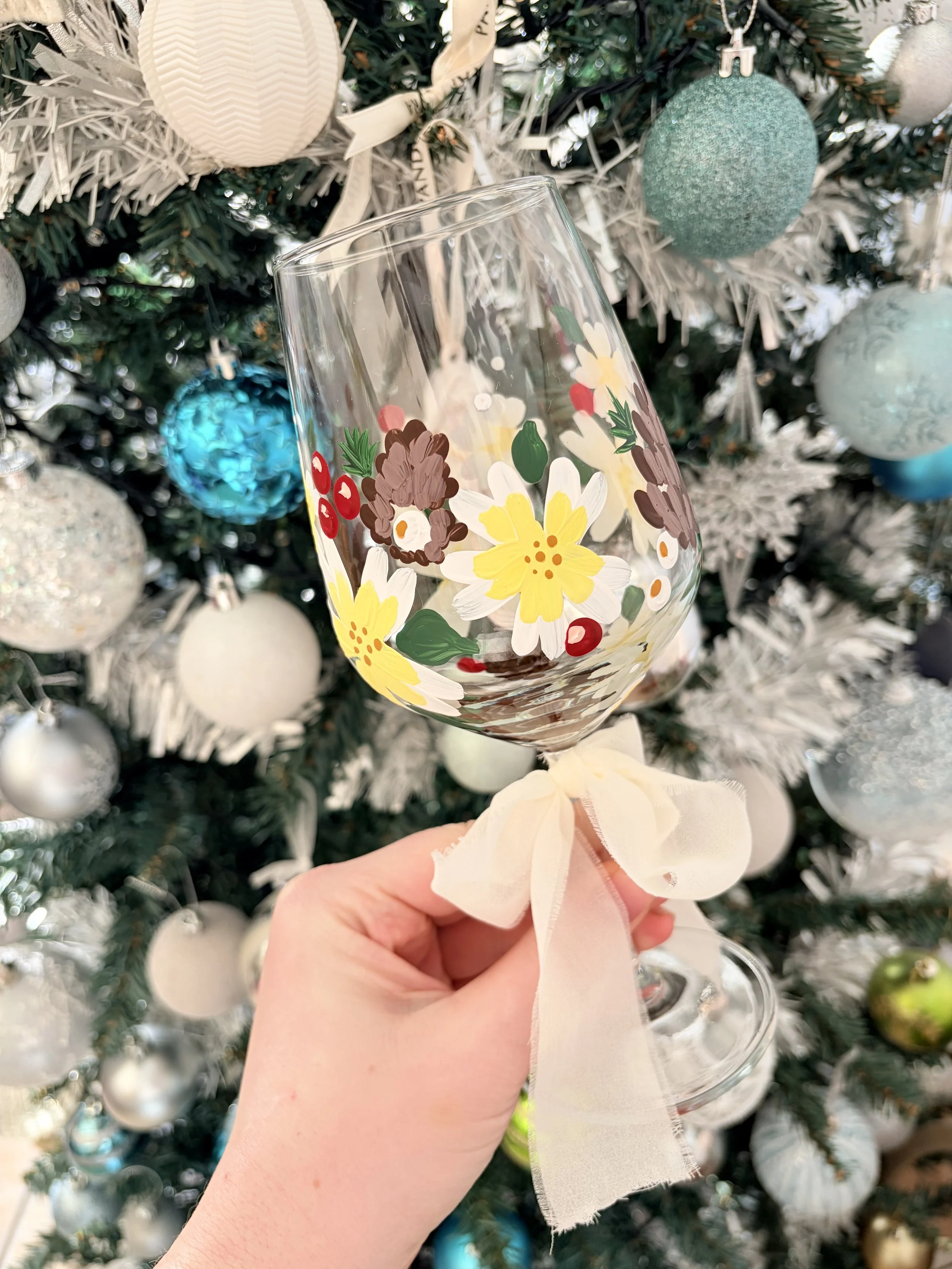 A hand holding a decorated wine glass with a white ribbon tied around the stem, in front of a Christmas tree decorated with white, silver, and blue ornaments.