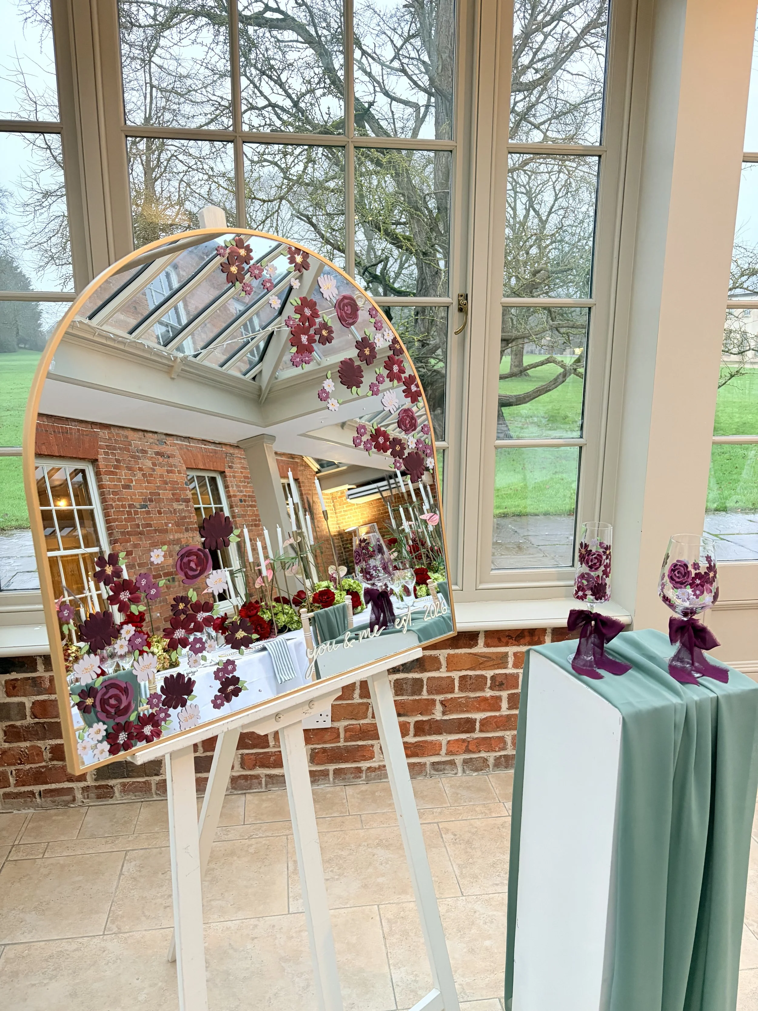 Decorative mirror with floral design, a table with glasses wrapped with purple ribbons, and a window showing an outdoor view with trees and grass.
