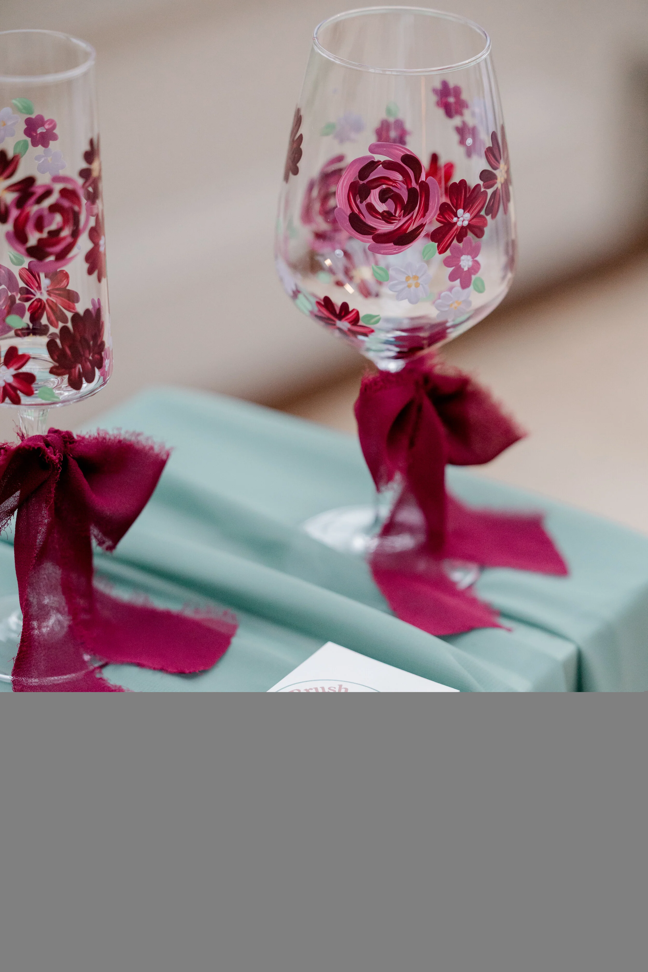 Two decorated wine glasses with flower patterns and pink ribbons, placed on a light blue gift box.