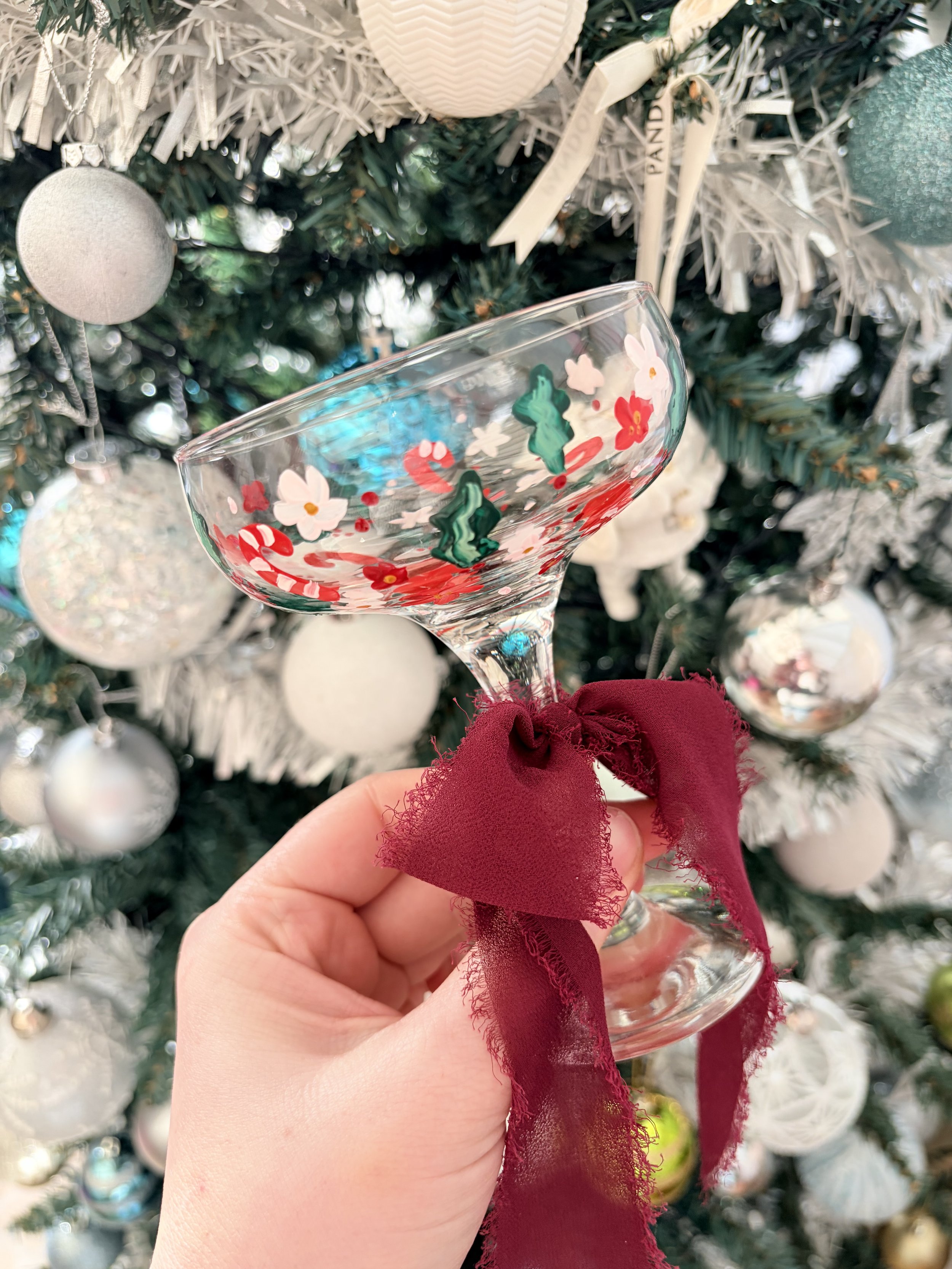 Hand holding a decorated glass with a red ribbon tied around it, in front of a decorated Christmas tree with white ornaments.