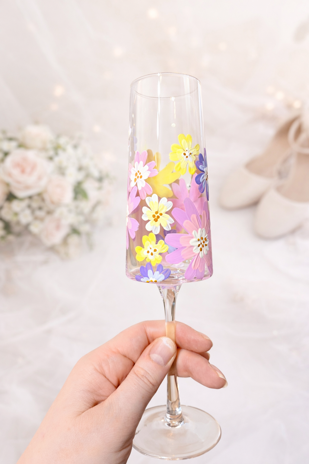 A hand holding a champagne flute decorated with colorful floral patterns, with a blurred background of flowers and shoes.