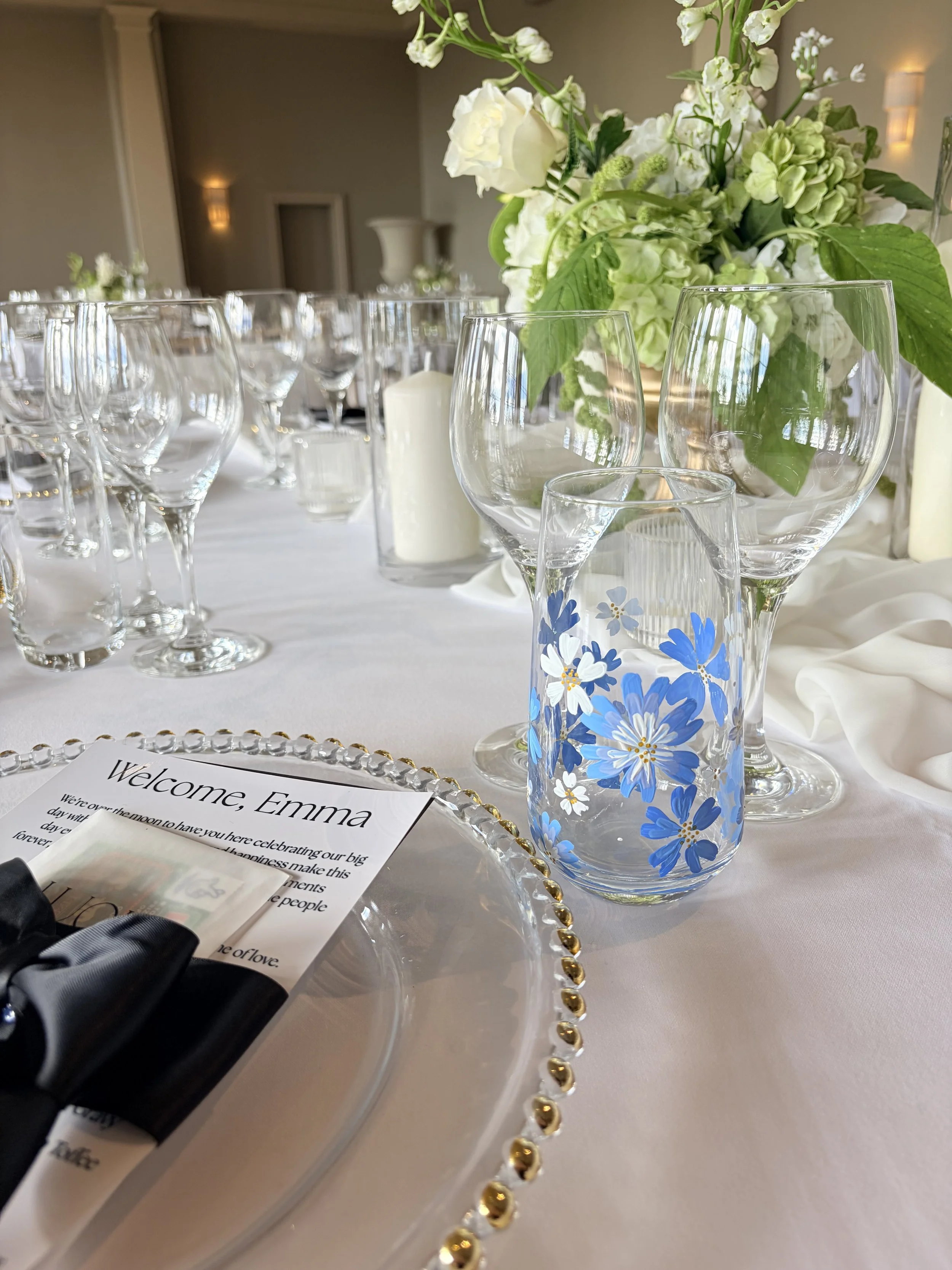 Elegant table setting with clear glassware, a floral centerpiece of white and green flowers, a white candle, and a welcome note on a decorative plate with gold trim, at a wedding reception.