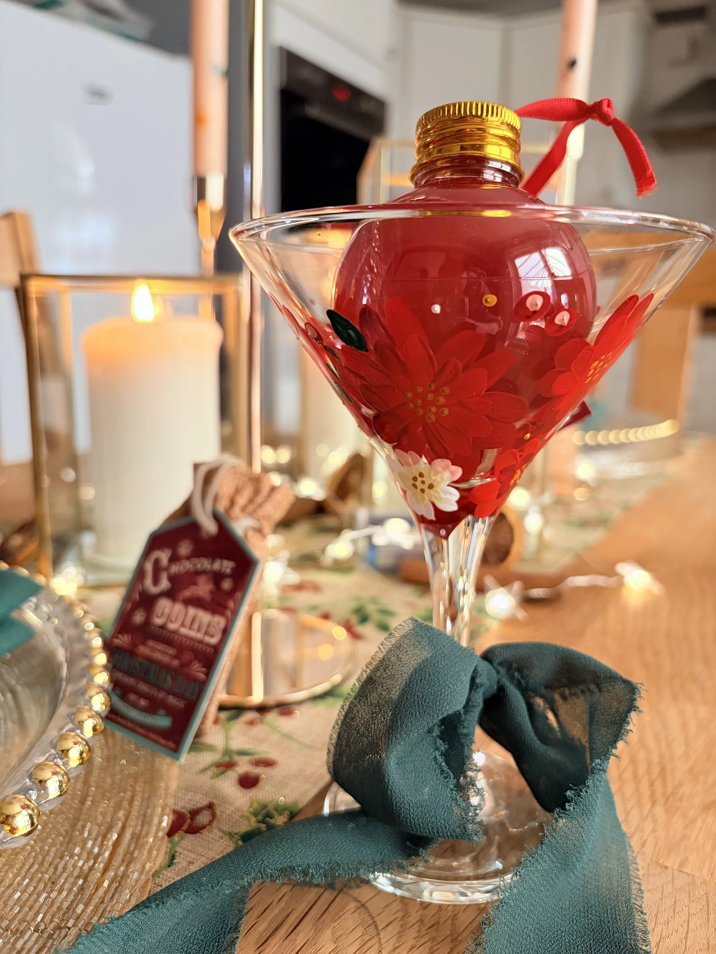 Decorative martini glass with red and white flowers painted on it, filled with red liquid, tied with a red ribbon, placed on a wooden table with holiday decorations.