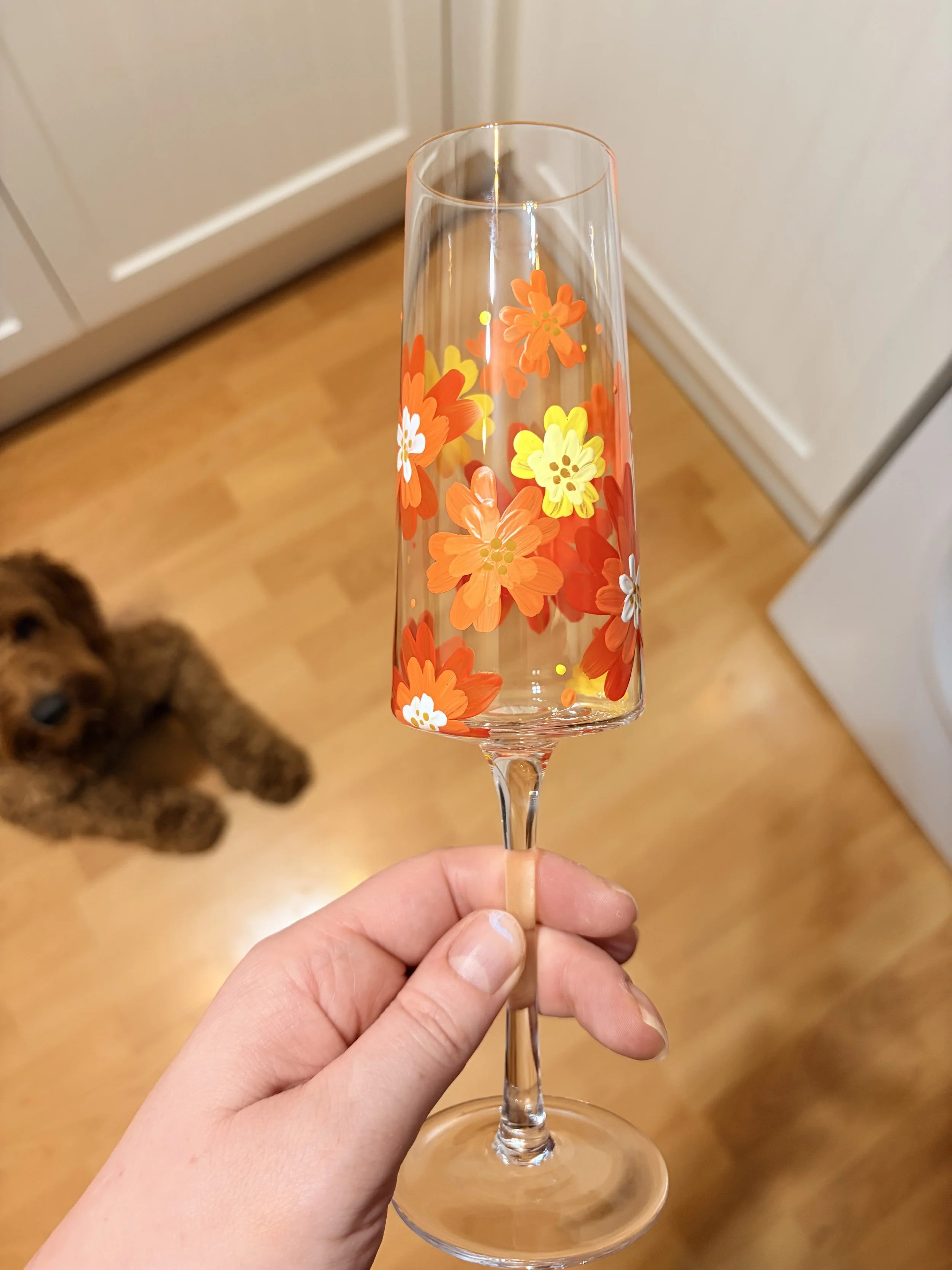 A hand holding a tall, narrow champagne flute decorated with colorful orange, yellow, white, and red floral patterns. A brown dog is visible in the background on a wooden floor.