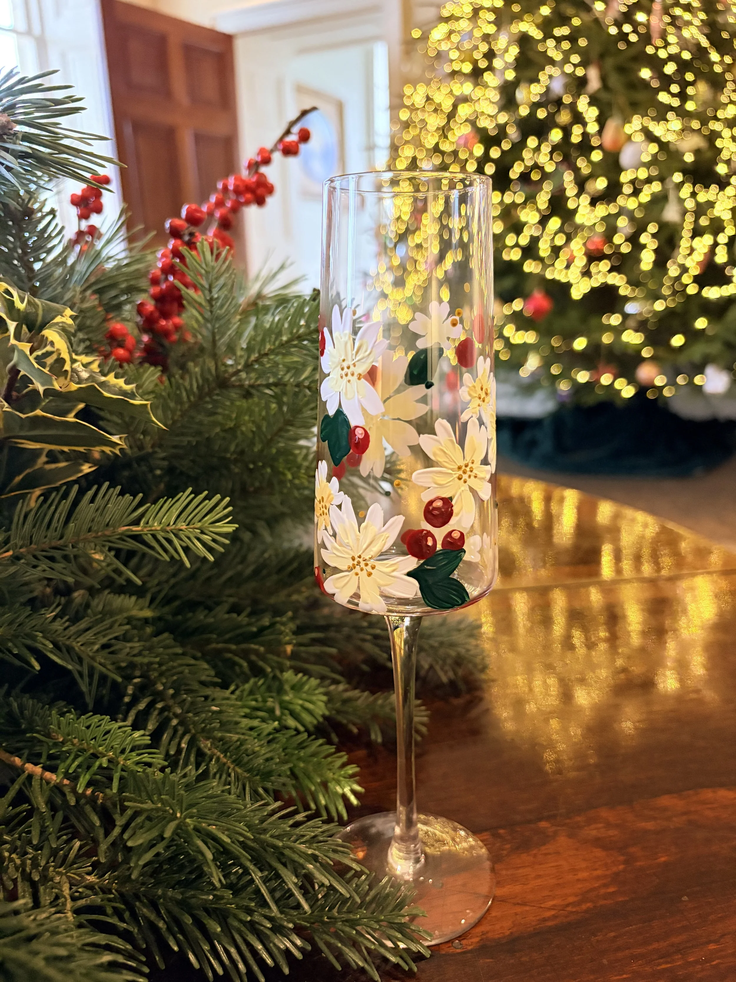 A decorated champagne flute with white flowers and red berries design, placed on a wooden table next to Christmas greenery with red berries, in front of a blurred Christmas tree with warm lights.