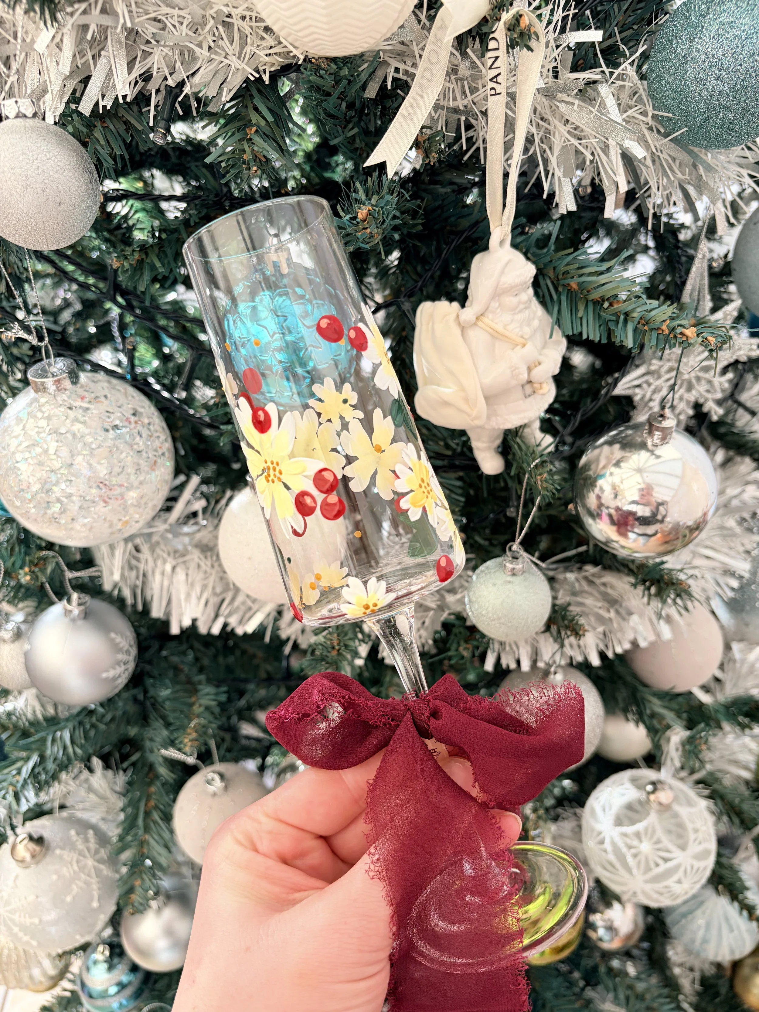 Hand holding a decorated champagne flute with red ribbon, in front of a decorated Christmas tree with white ornaments and a Santa figurine ornament.