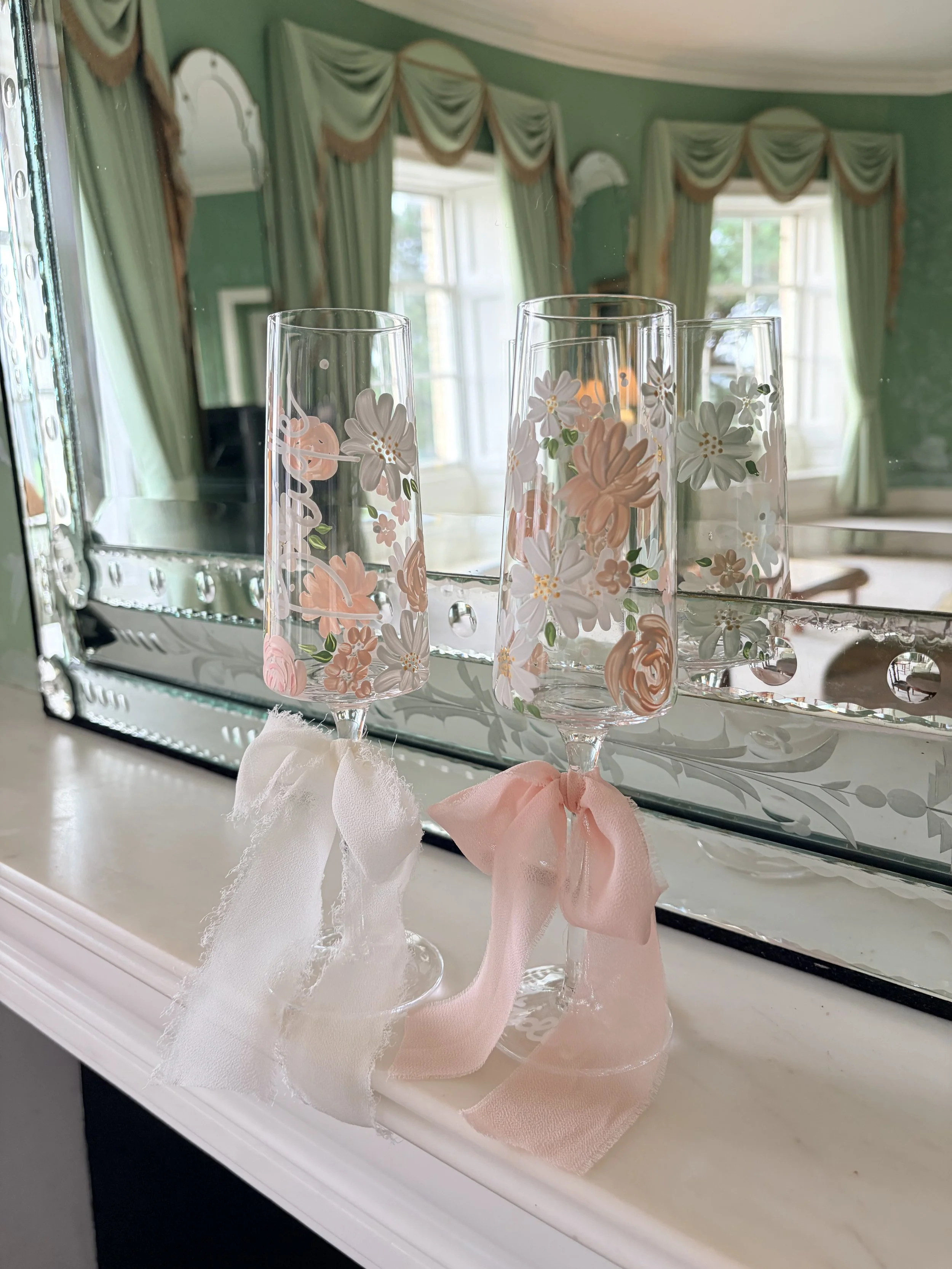 Three flute glasses with floral patterns and pink bows, placed in front of a mirror on a white surface in a green-colored room with large windows and draped curtains.