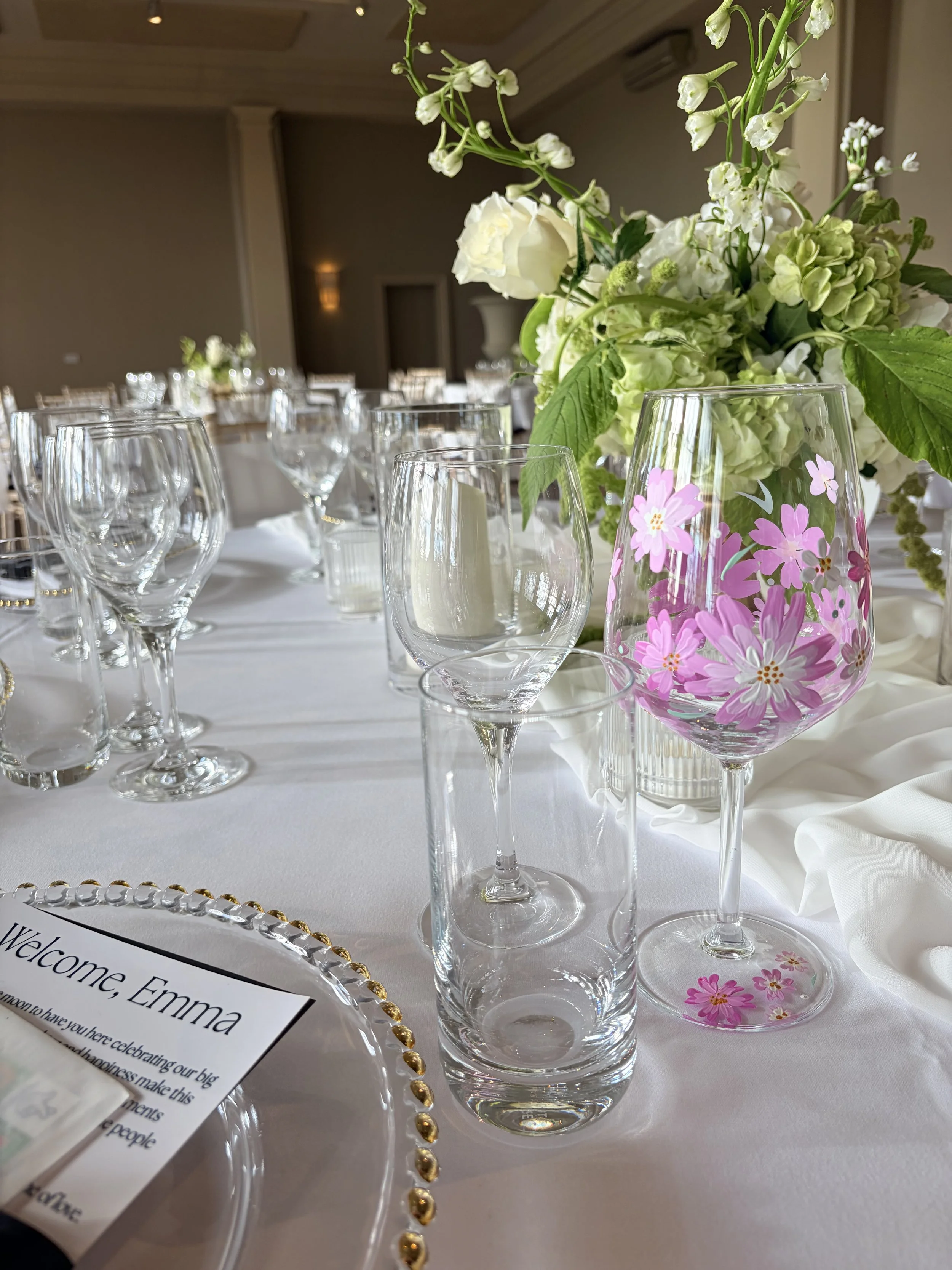 Elegant place setting at a wedding reception with a white tablecloth, various empty glassware, a floral centerpiece with white and green flowers, and a glass with pink and white painted flowers, alongside a welcome card for Emma.