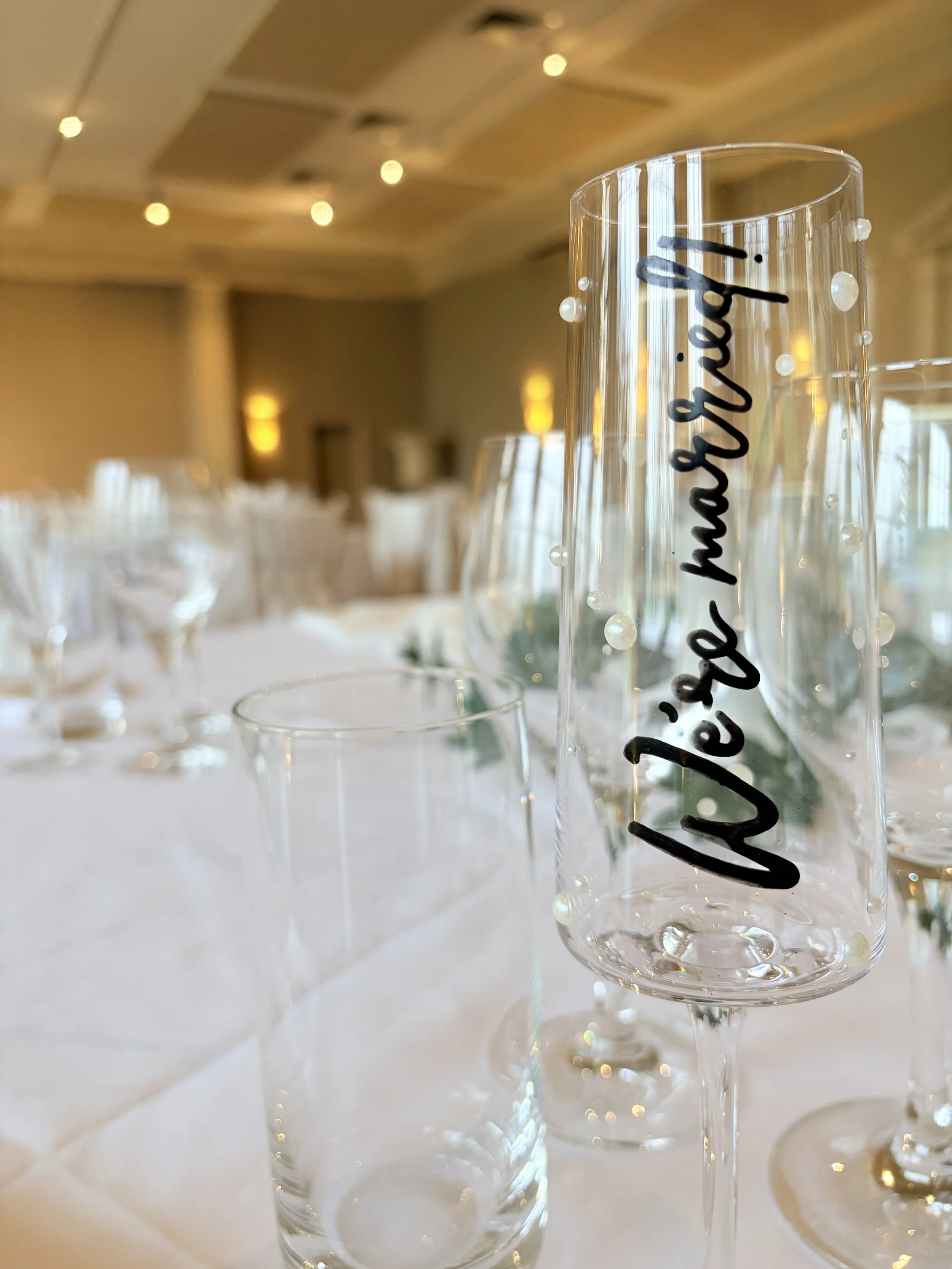 A close-up of a champagne flute with the phrase 'We're getting married' written on it, set on a decorated wedding reception table with more glasses and a blurred background of a banquet hall.