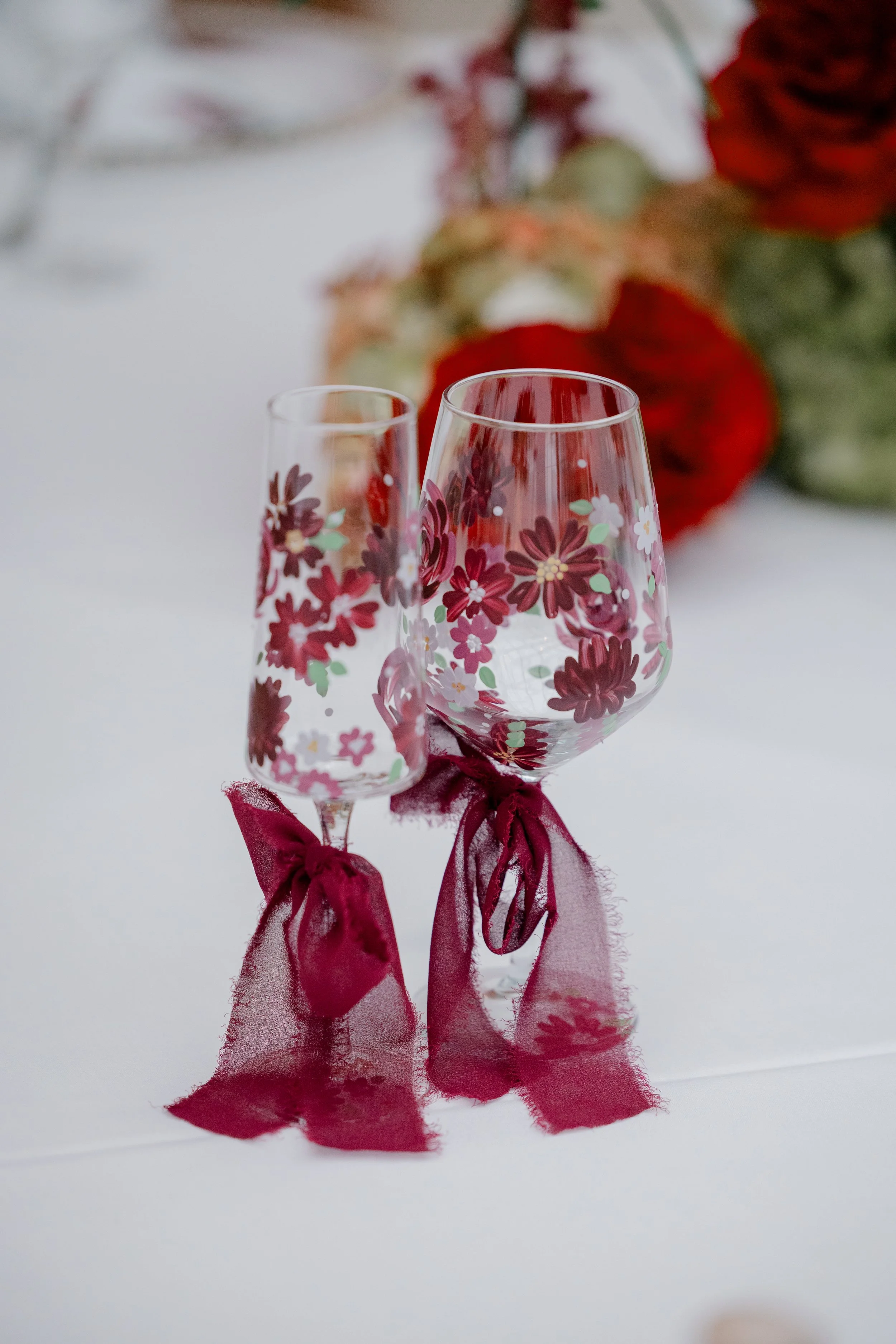 Two wine glasses decorated with red ribbons and floral patterns, set on a white tablecloth, with a blurred floral centerpiece in the background.