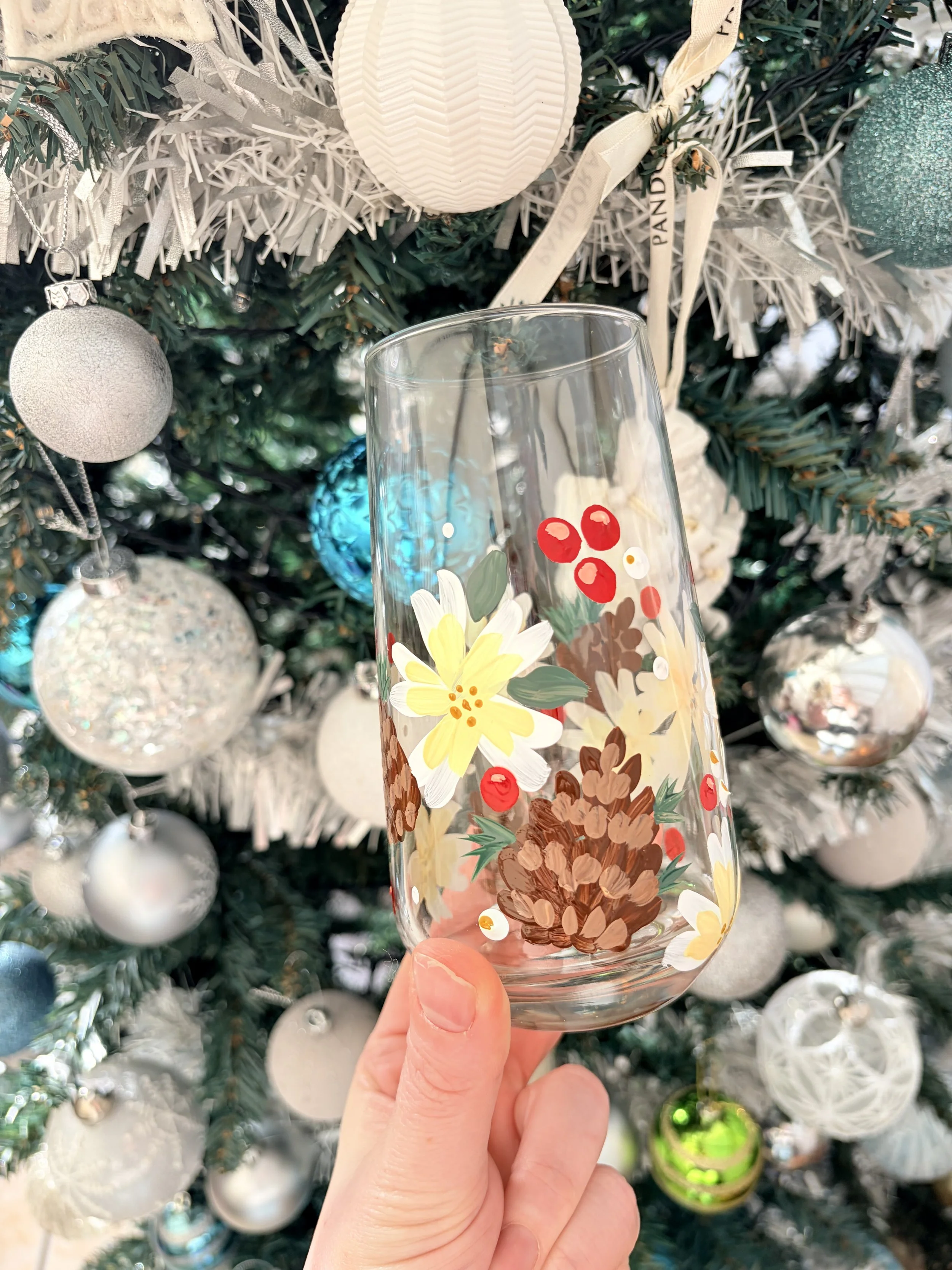 A clear glass with a painted floral design featuring pinecones, yellow flowers, red berries, and green leaves, held in front of a Christmas tree decorated with various white, silver, blue, and green ornaments.