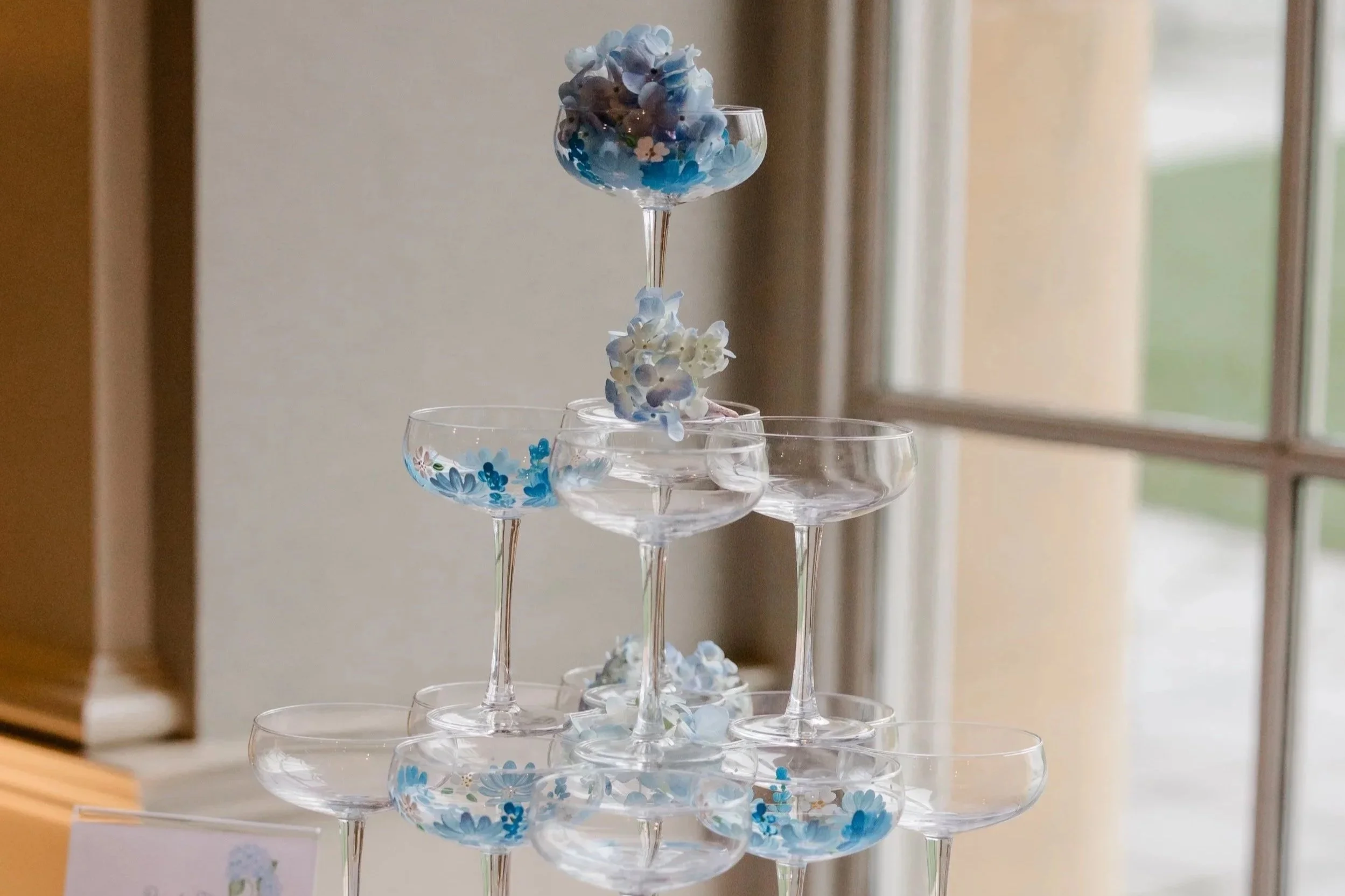 A pyramid of clear cocktail glasses decorated with blue, white, and purple floral designs, with some flowers inside the glasses, set near a window.