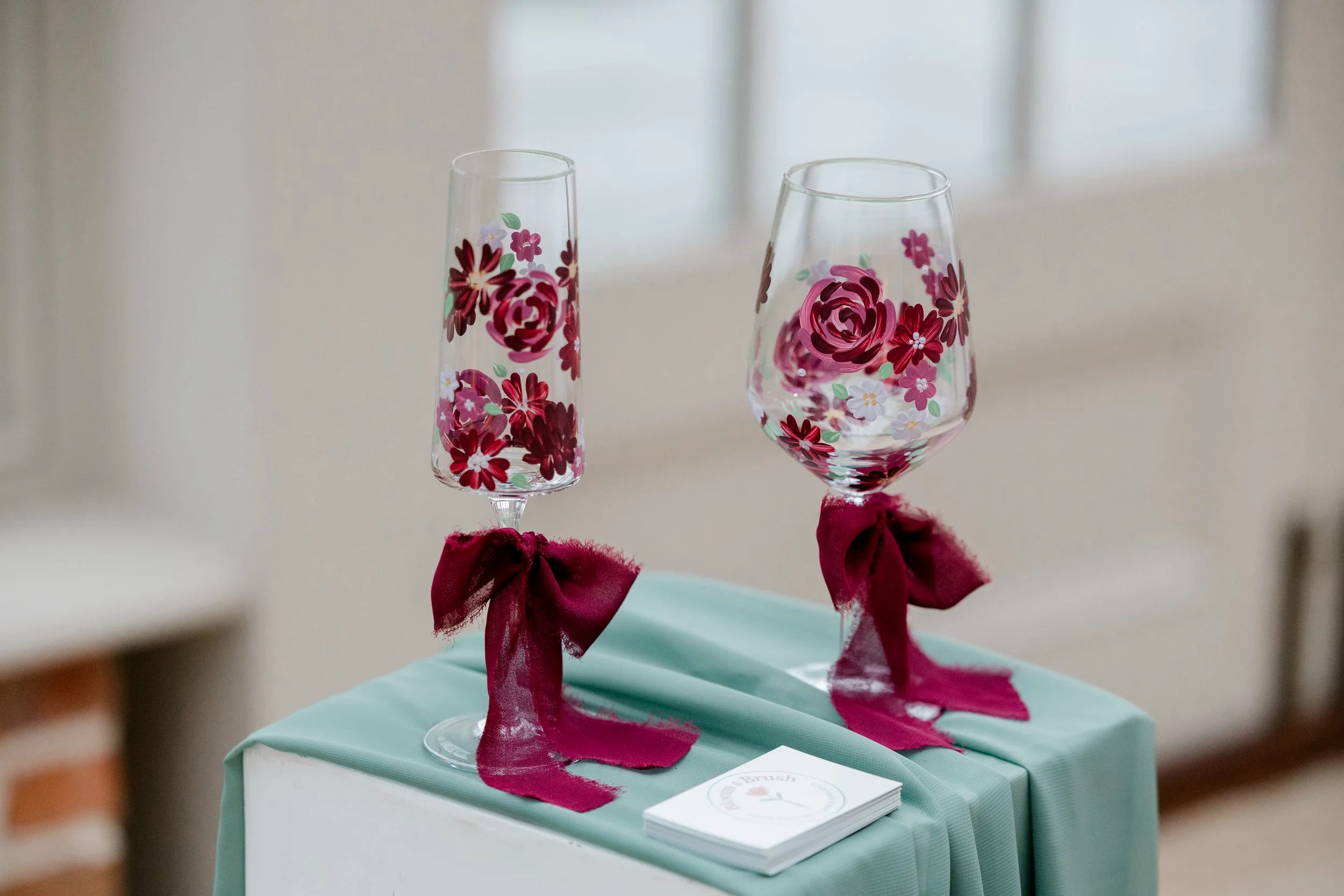 Two decorated wine glasses with red and pink floral patterns, tied with red ribbons, placed on a table with a light blue tablecloth.