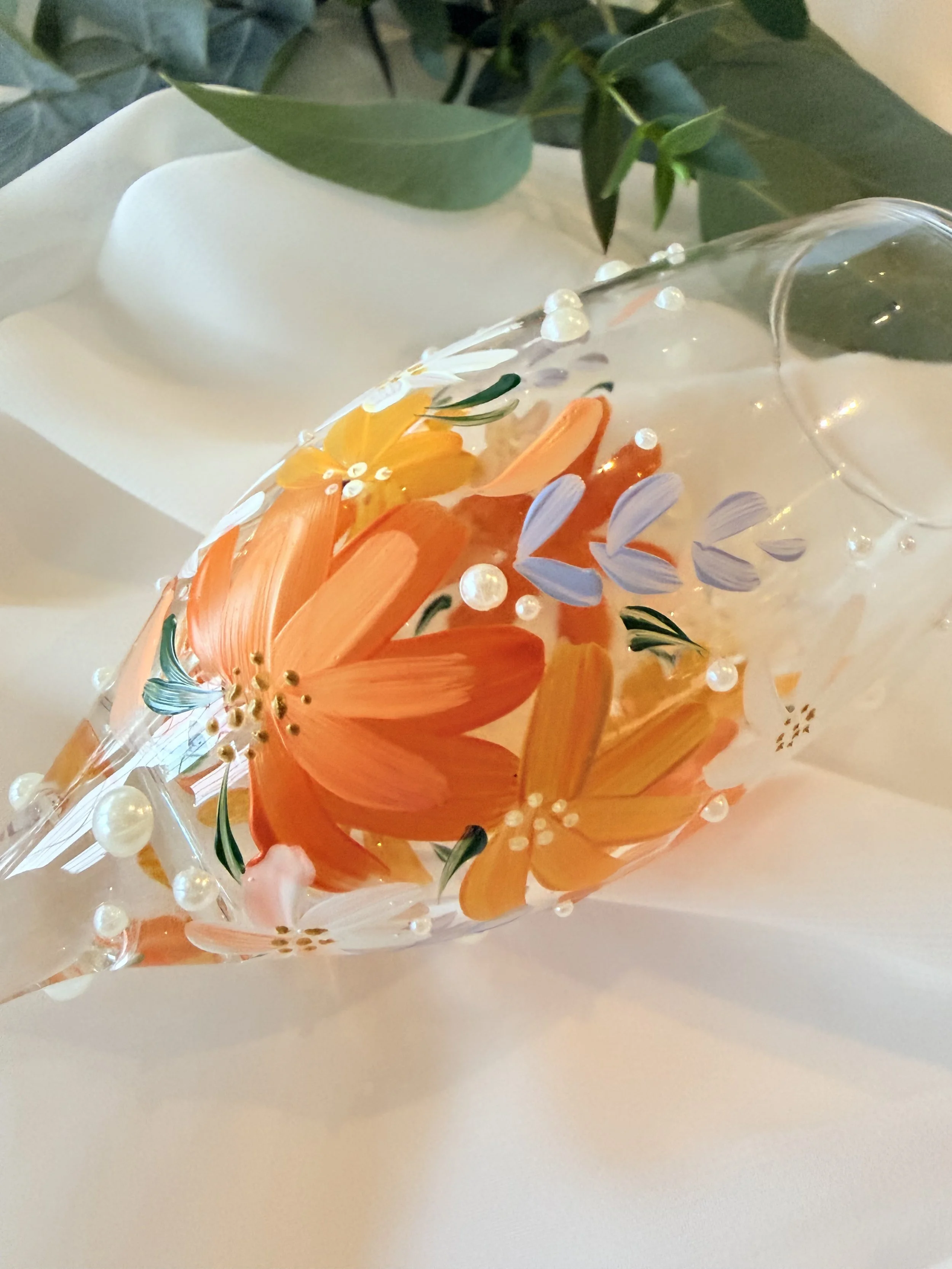 Decorative glass with painted floral design containing orange, yellow, and purple flowers, surrounded by green leaves and small white beads, on a white surface with green leaves in the background.
