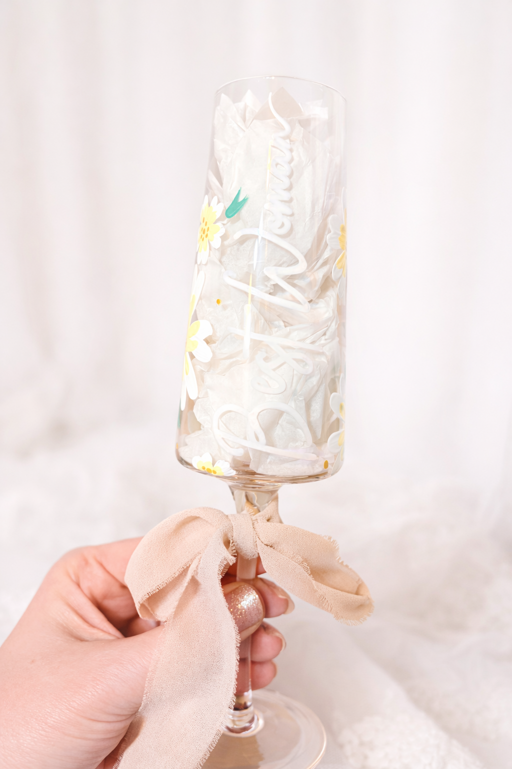 A hand holding a champagne flute decorated with a pink ribbon, filled with crumpled tissue paper, with a white background.