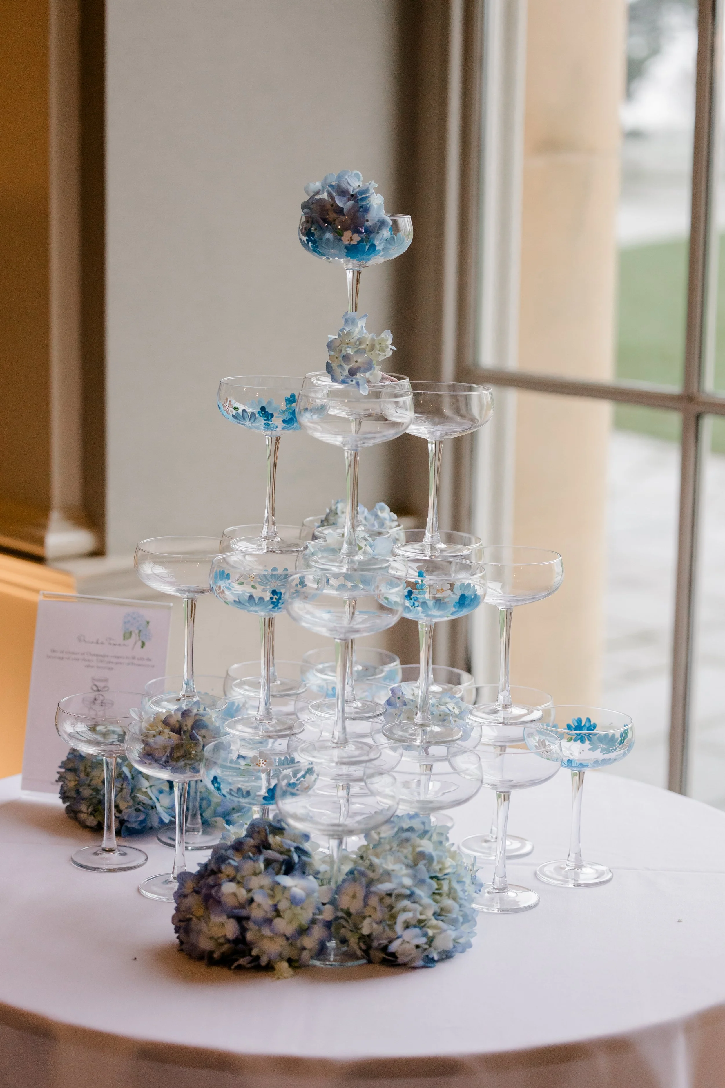 A pyramid of empty champagne glasses decorated with blue and white hydrangea flowers, with more hydrangeas at the base, set on a round table near a window.